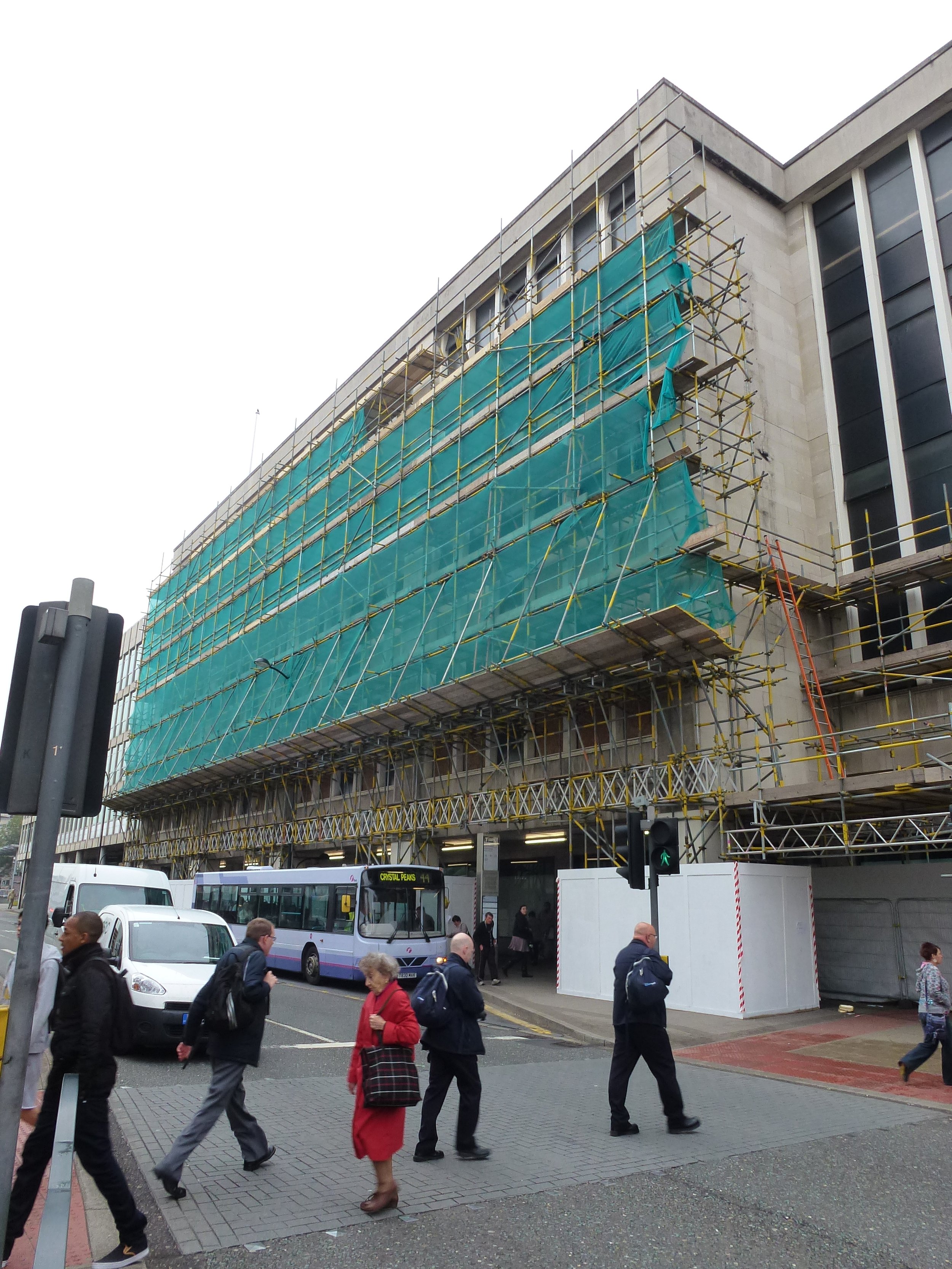 People walking on a city street in front of a large building under construction with scaffolding and green safety nets.