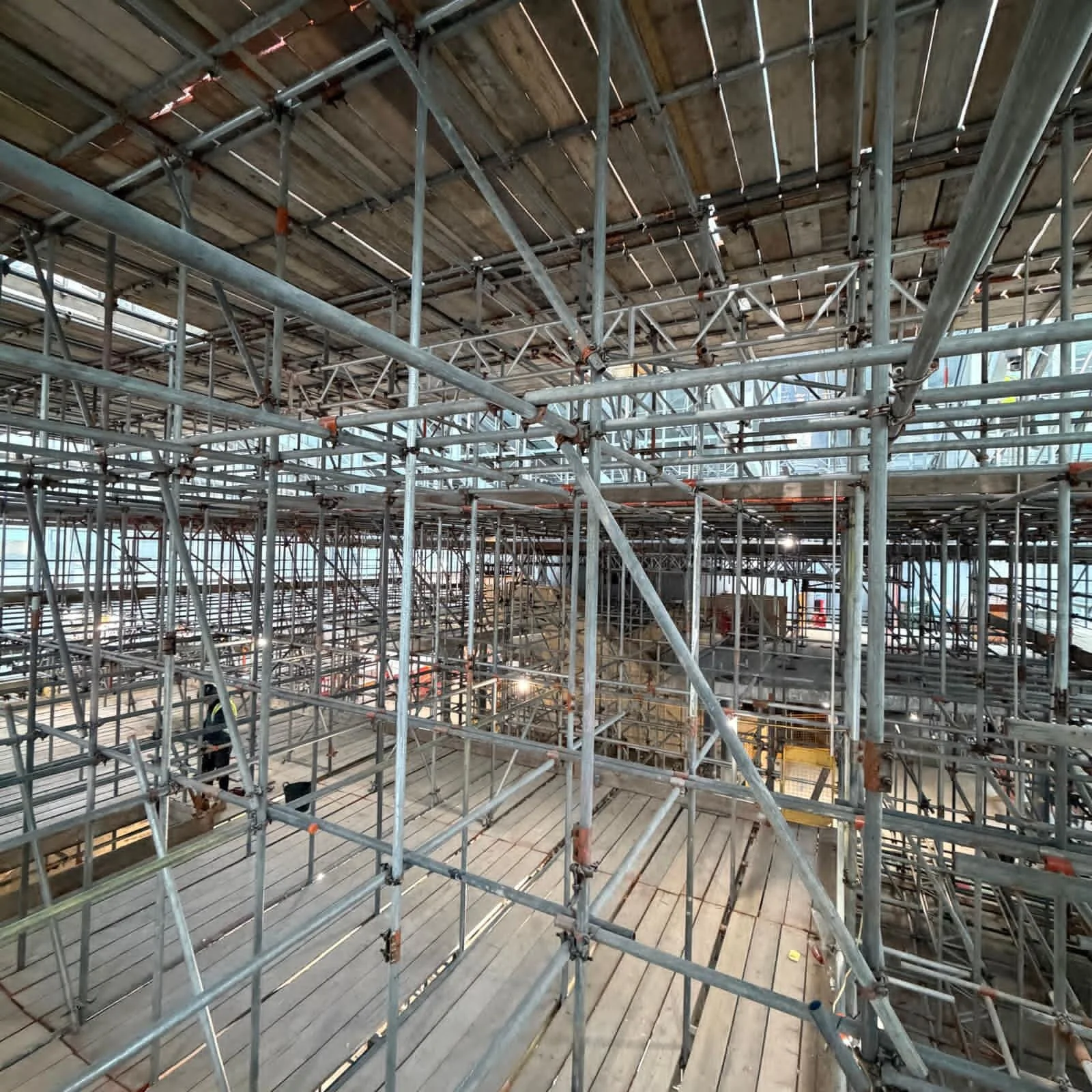 Interior view of a construction site with extensive metal scaffolding and wooden flooring, indicating ongoing building work in a large structure.