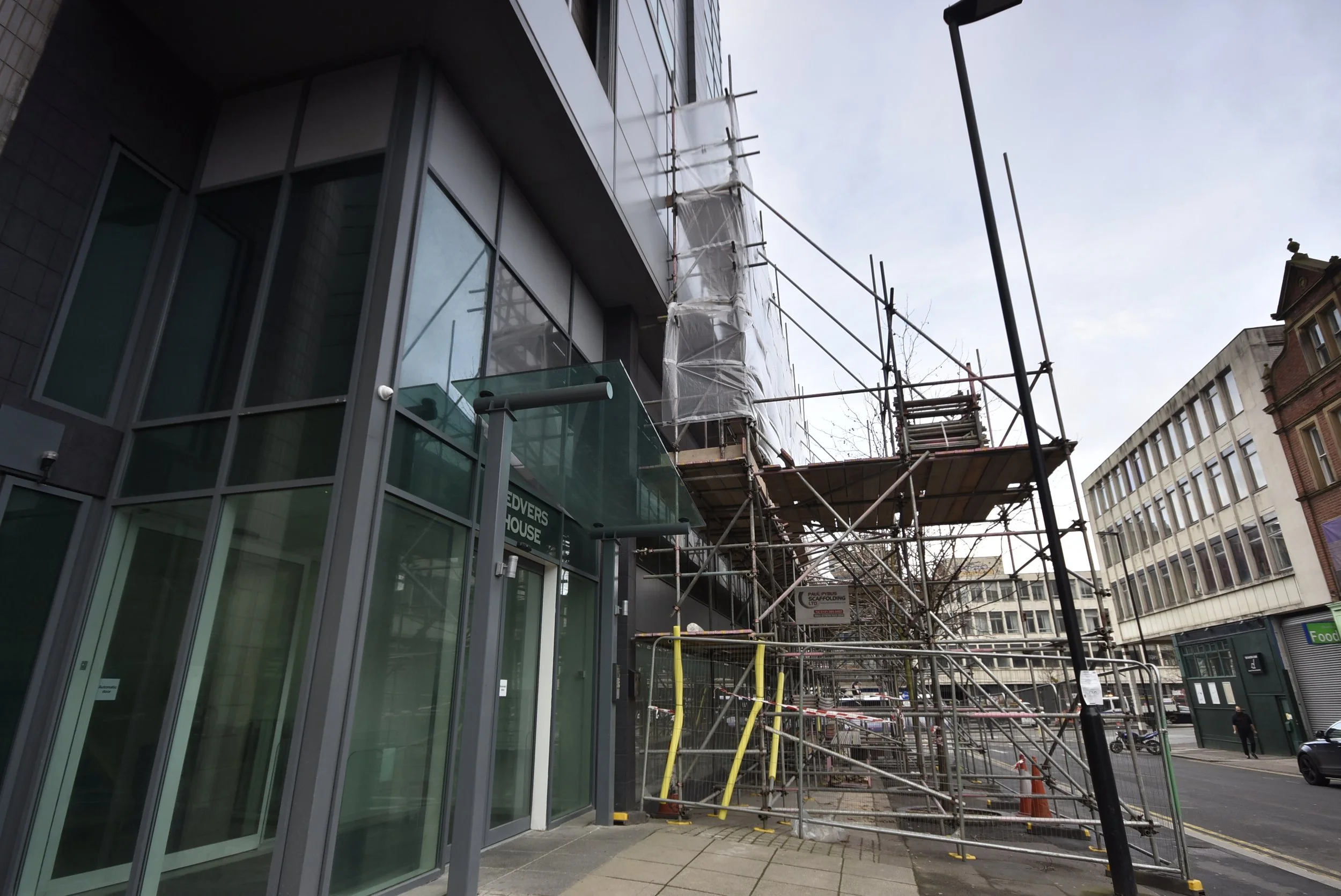 Construction scaffolding surrounds a modern glass-front building on an urban street, with a passerby walking in the background.