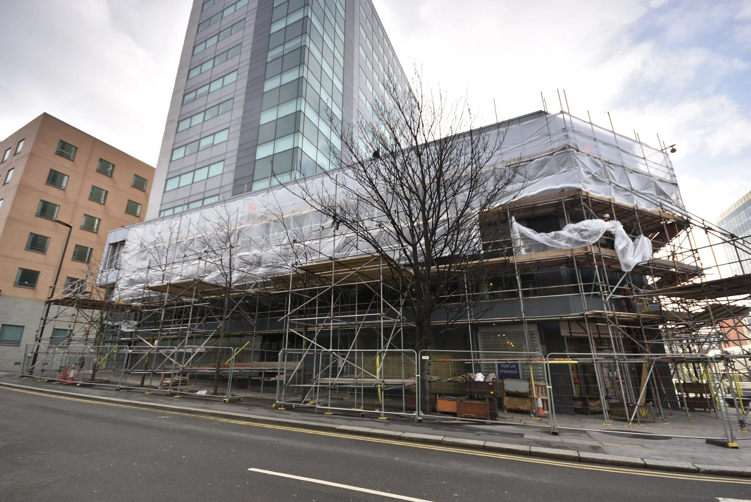 Street view of a building under construction with scaffolding and white protective sheeting, surrounded by a metal fence, with a bare tree in the foreground and modern high-rise buildings in the background.