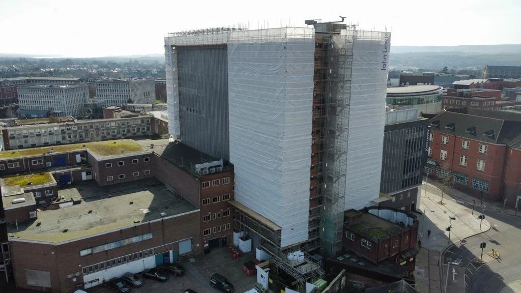 A large building under construction wrapped in white protective covering, with scaffolding and a construction elevator on the side. The surrounding area includes other brick and modern buildings and a street with cars and pedestrians.