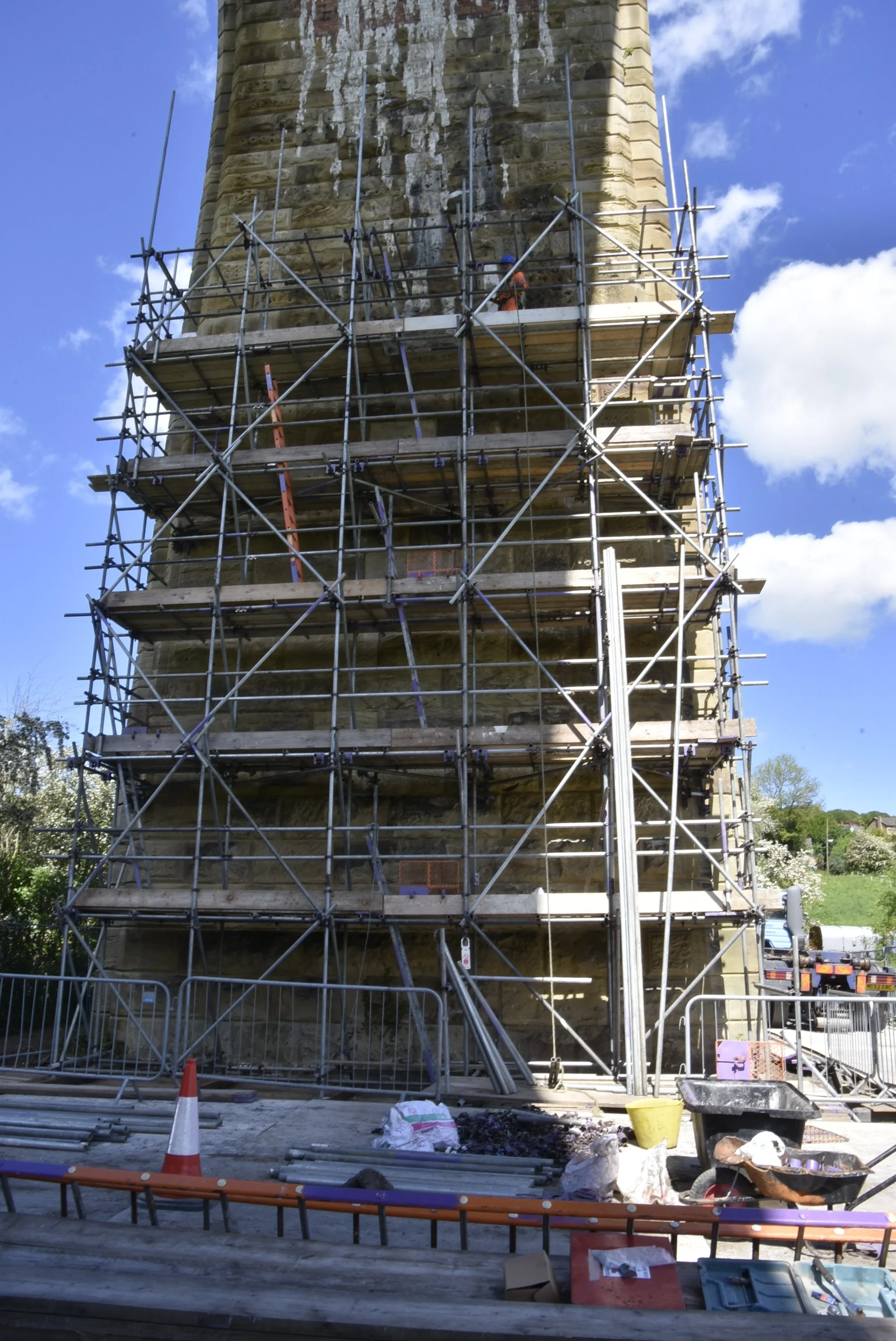 A tall stone tower under renovation with scaffolding around it. Small construction workers are visible on the scaffolding. The sky is blue with some white clouds.