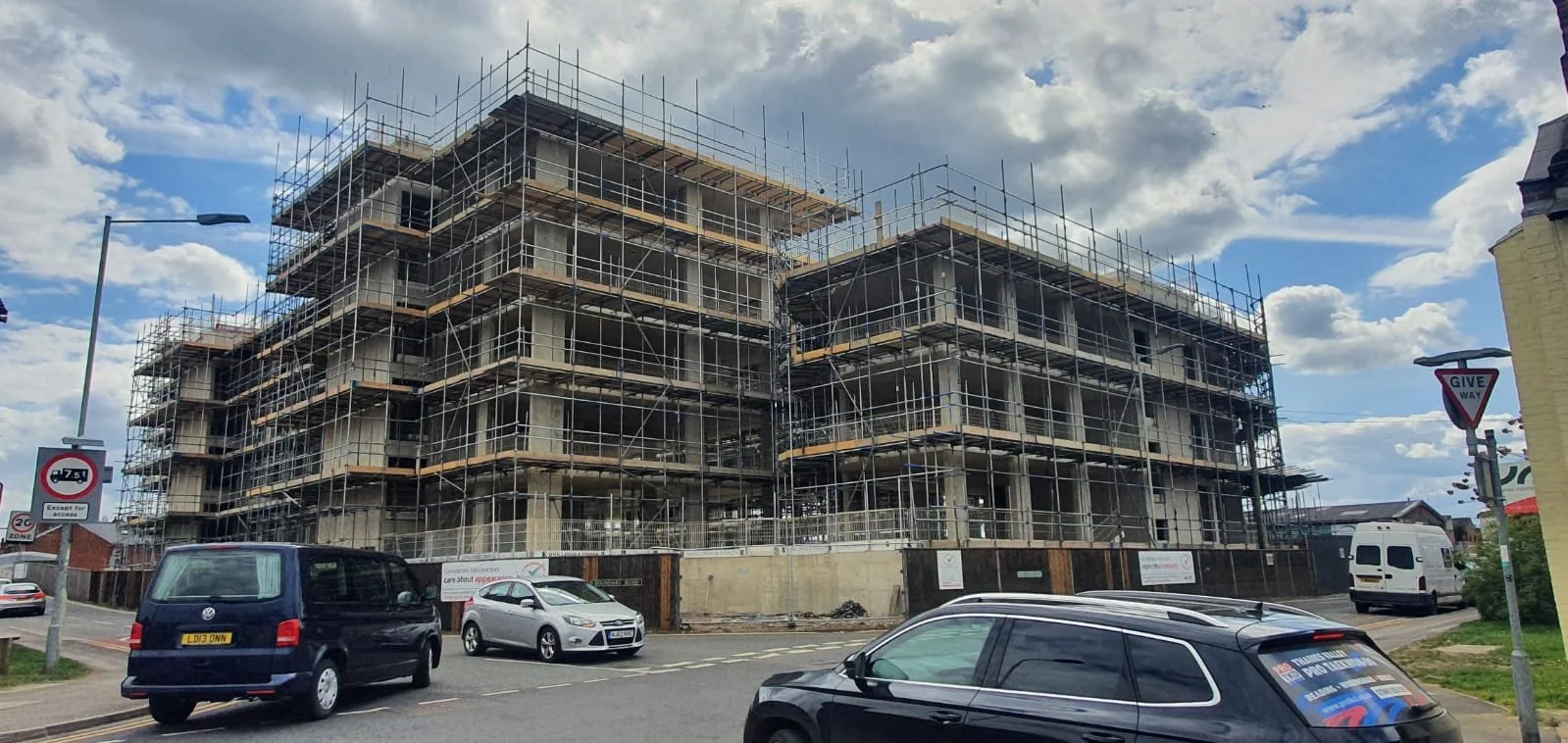 Construction site with multi-story building under construction, surrounded by scaffolding, near parked cars and street signs under a partly cloudy sky.