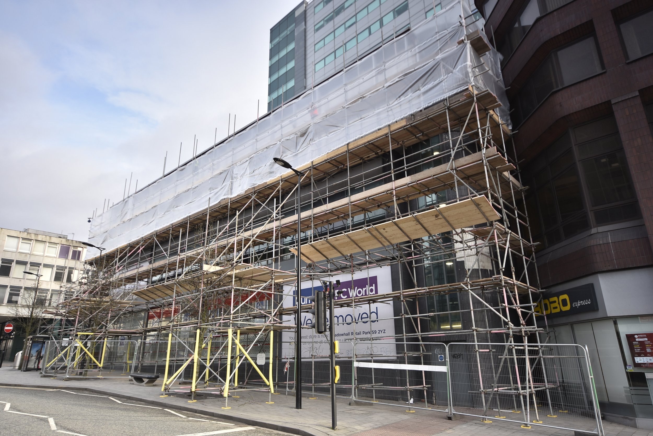 Scaffolding on a city building under construction, with protective sheeting and signage for a store, alongside streetlights and barriers.