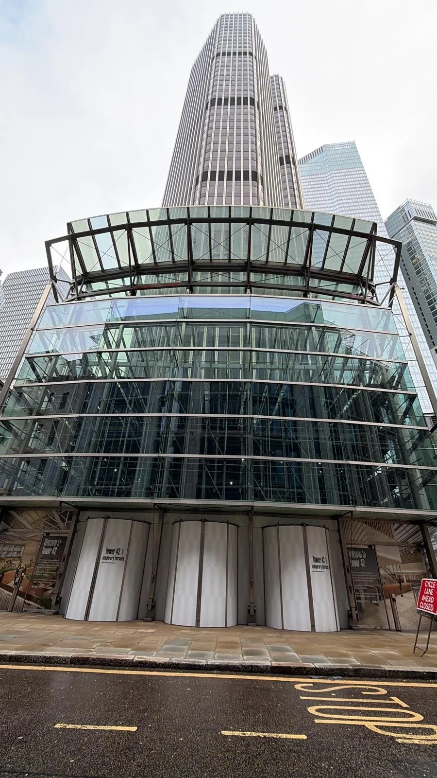 A modern glass skyscraper, known as Tower 42, in an urban cityscape with other high-rise buildings around it.