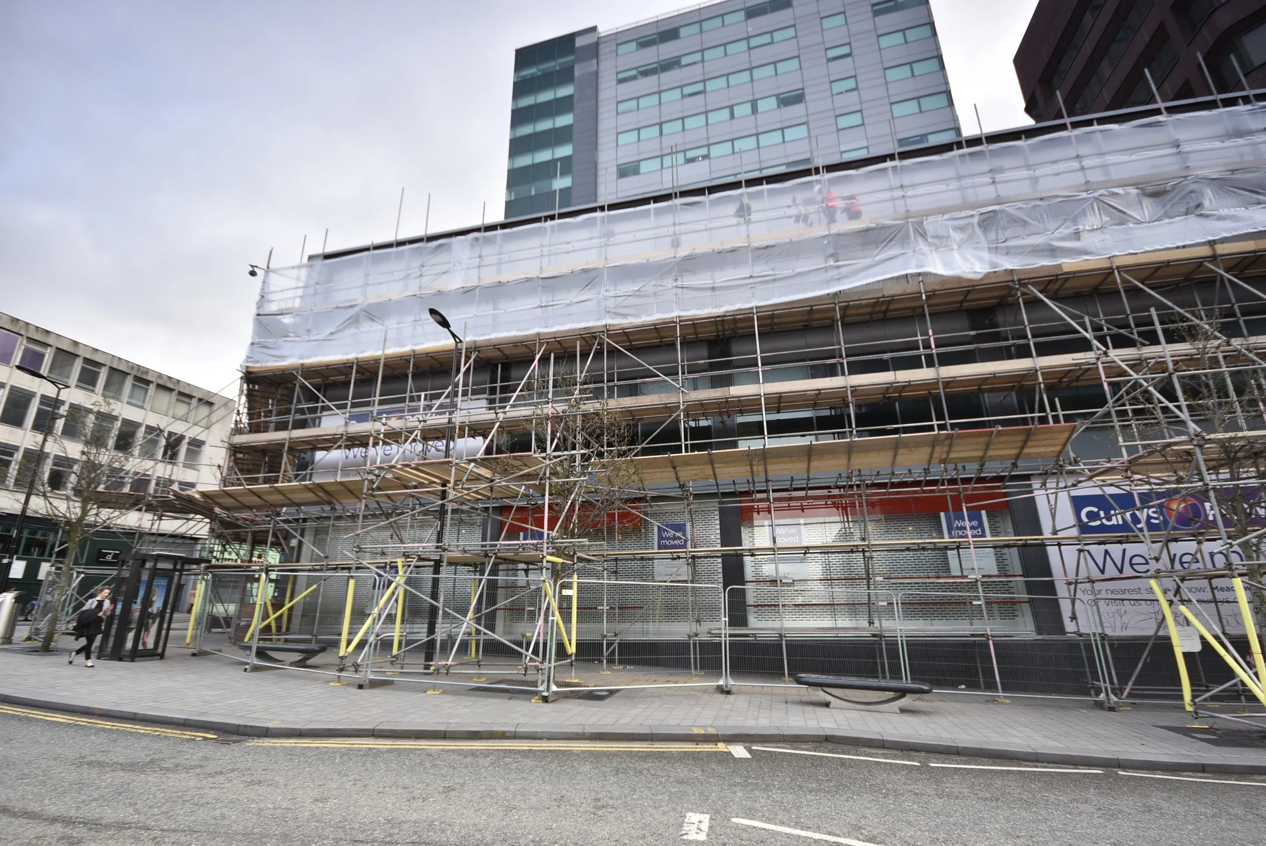Building under construction with scaffolding on a city street, modern high-rise buildings in the background, and a person walking near a bus stop.