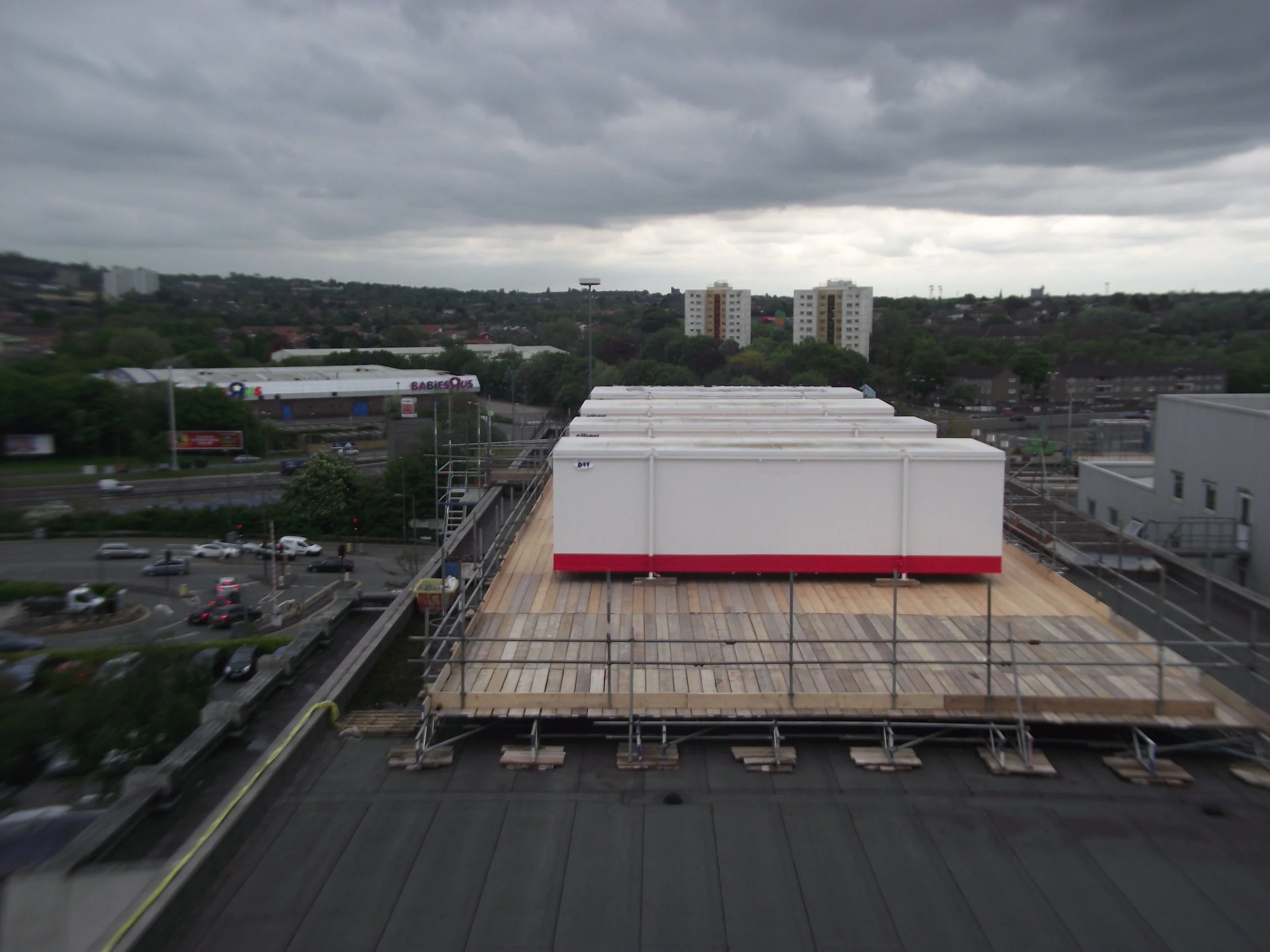 Rooftop under construction with white modular units on a wooden platform, surrounded by scaffolding, overlooking a busy street and residential buildings under a cloudy sky.
