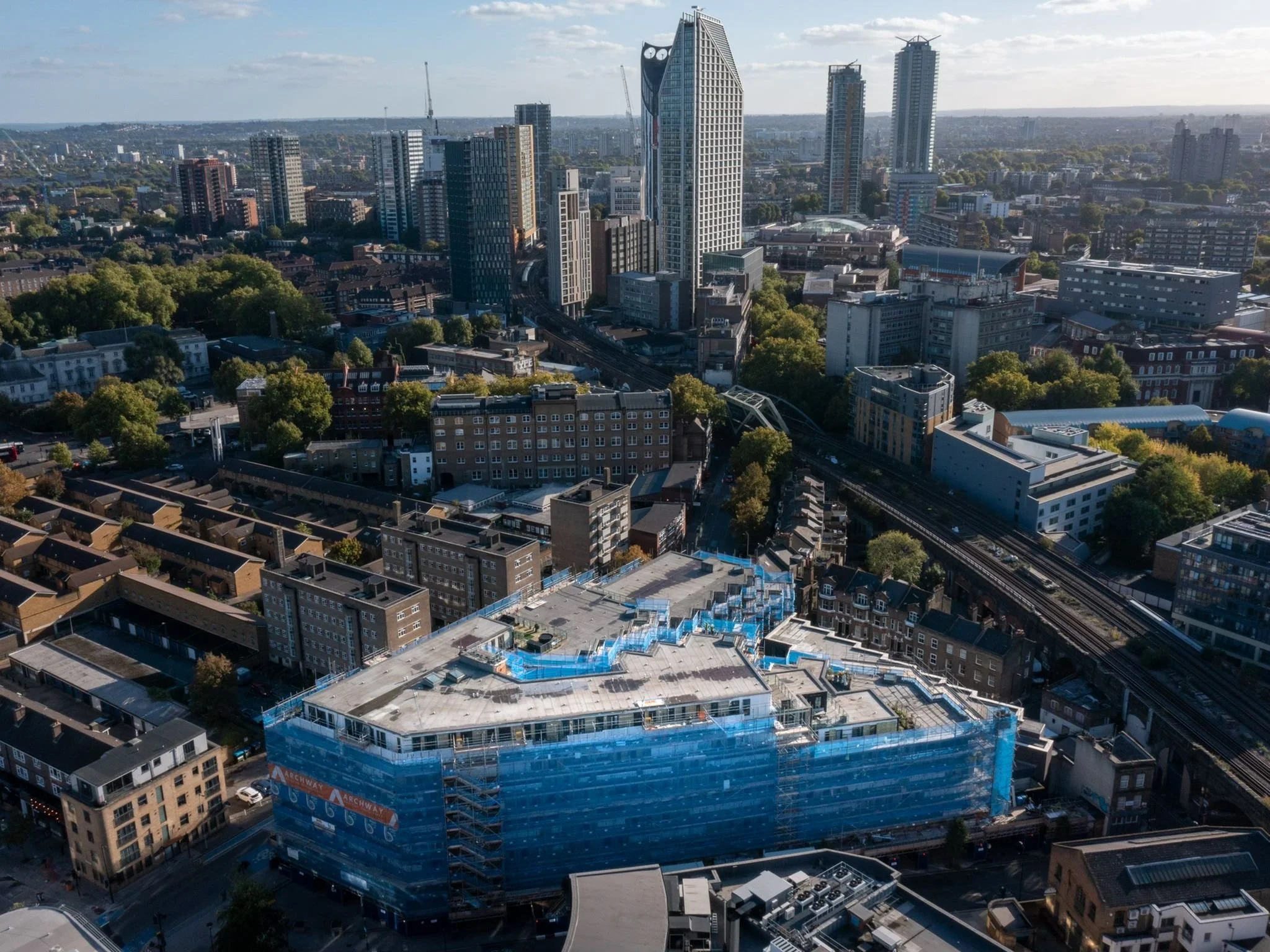 A cityscape with tall modern buildings, some still under construction, surrounded by greenery. A commercial building covered in blue construction netting is visible in the foreground.