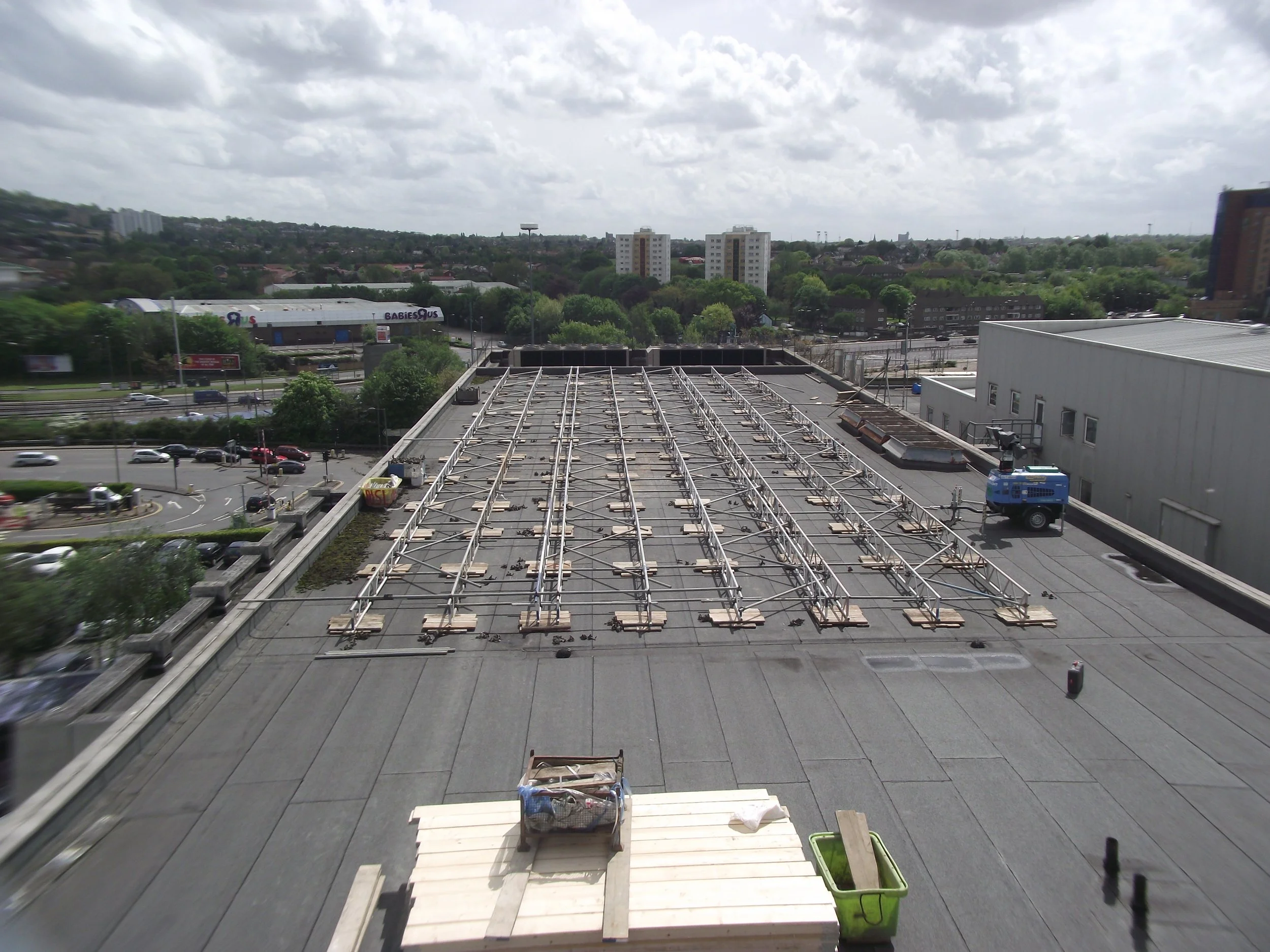 View of a rooftop with metal framing structures and construction tools, against a background of a cityscape with buildings, trees, and a cloudy sky.