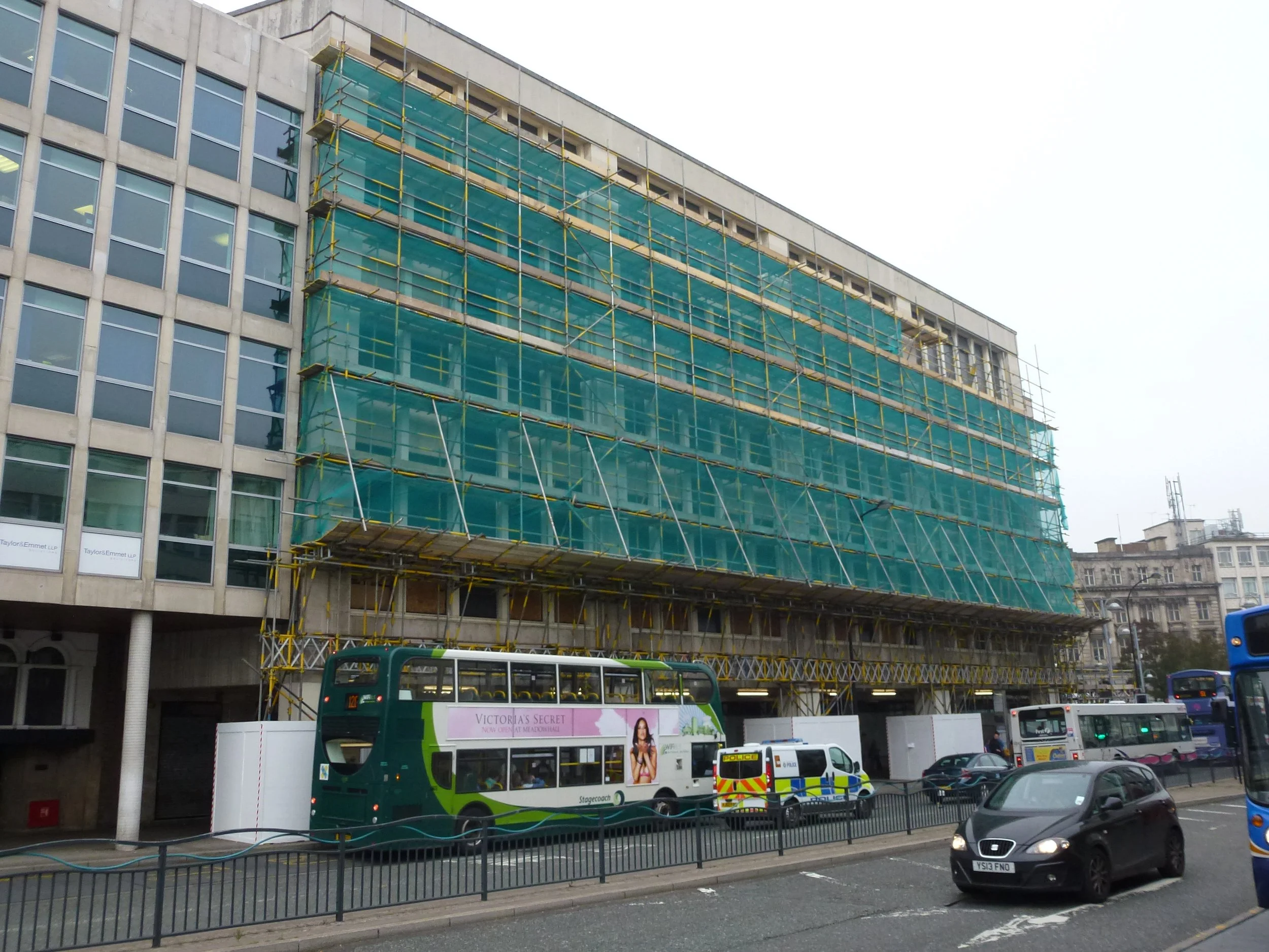 Street scene with a building under construction covered in green scaffolding, a double-decker bus with an advertisement, police vehicle, and cars and buses on the road.