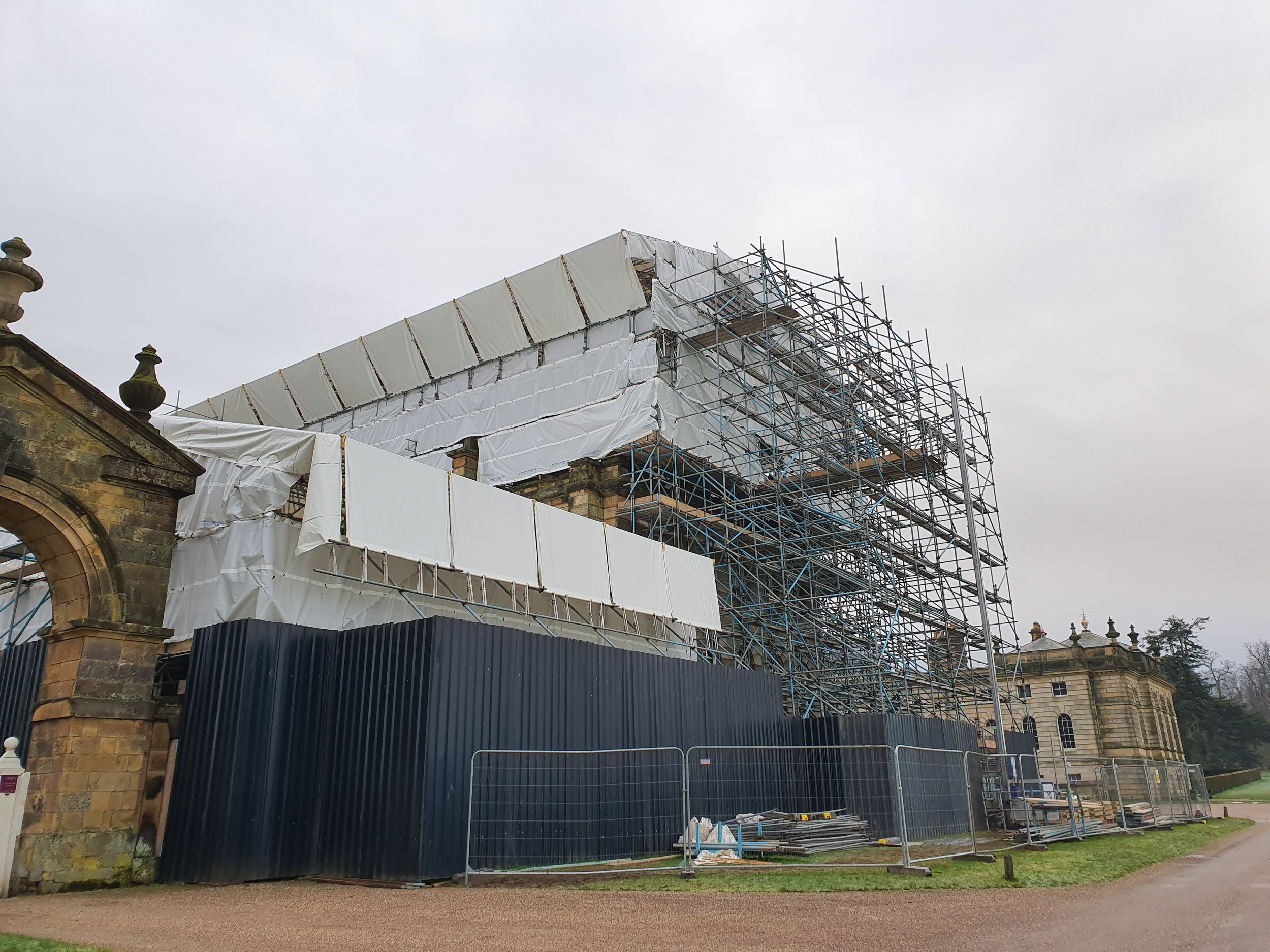 A historic building undergoing renovation with scaffolding and protective coverings, surrounded by fencing.
