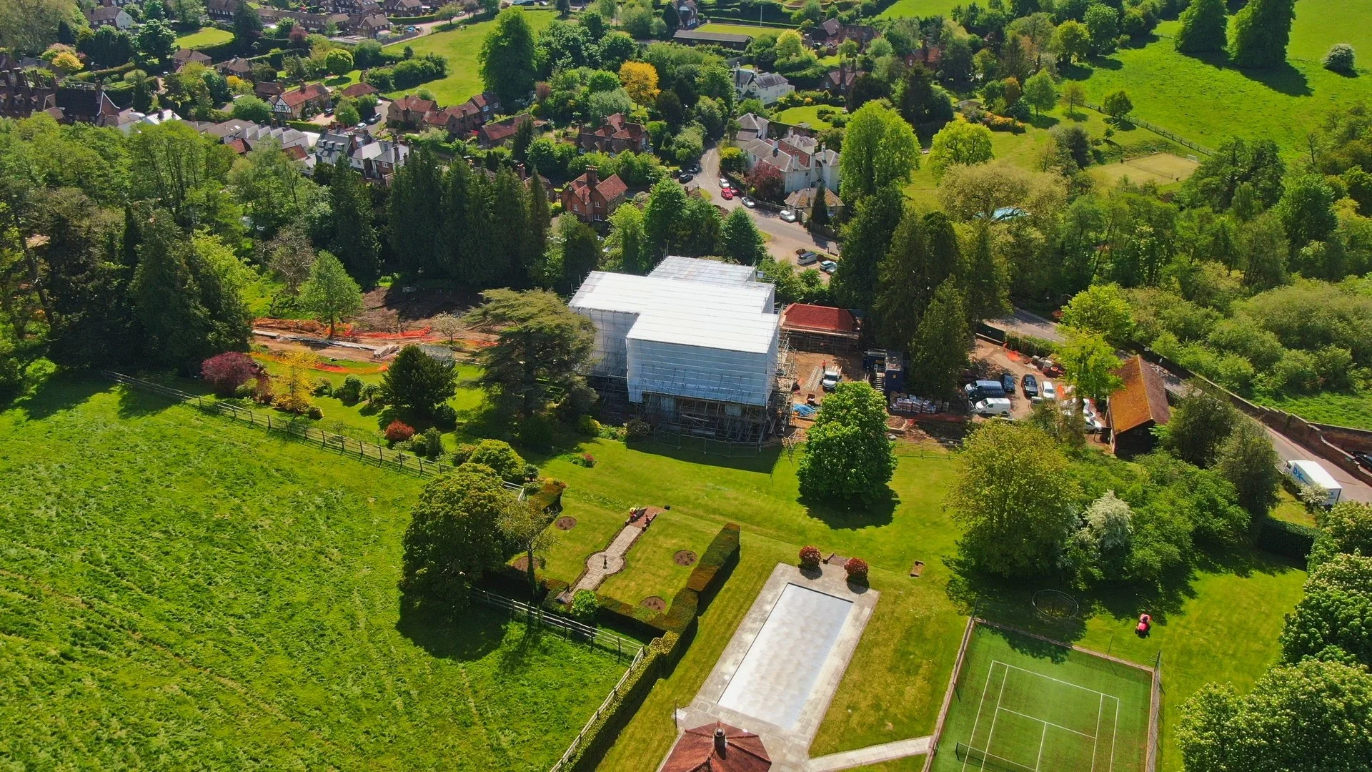An aerial view of a large residential backyard with a tennis court, green lawn, trees, and ongoing construction covered with scaffolding and a white tarp. Houses and greenery surround the area.