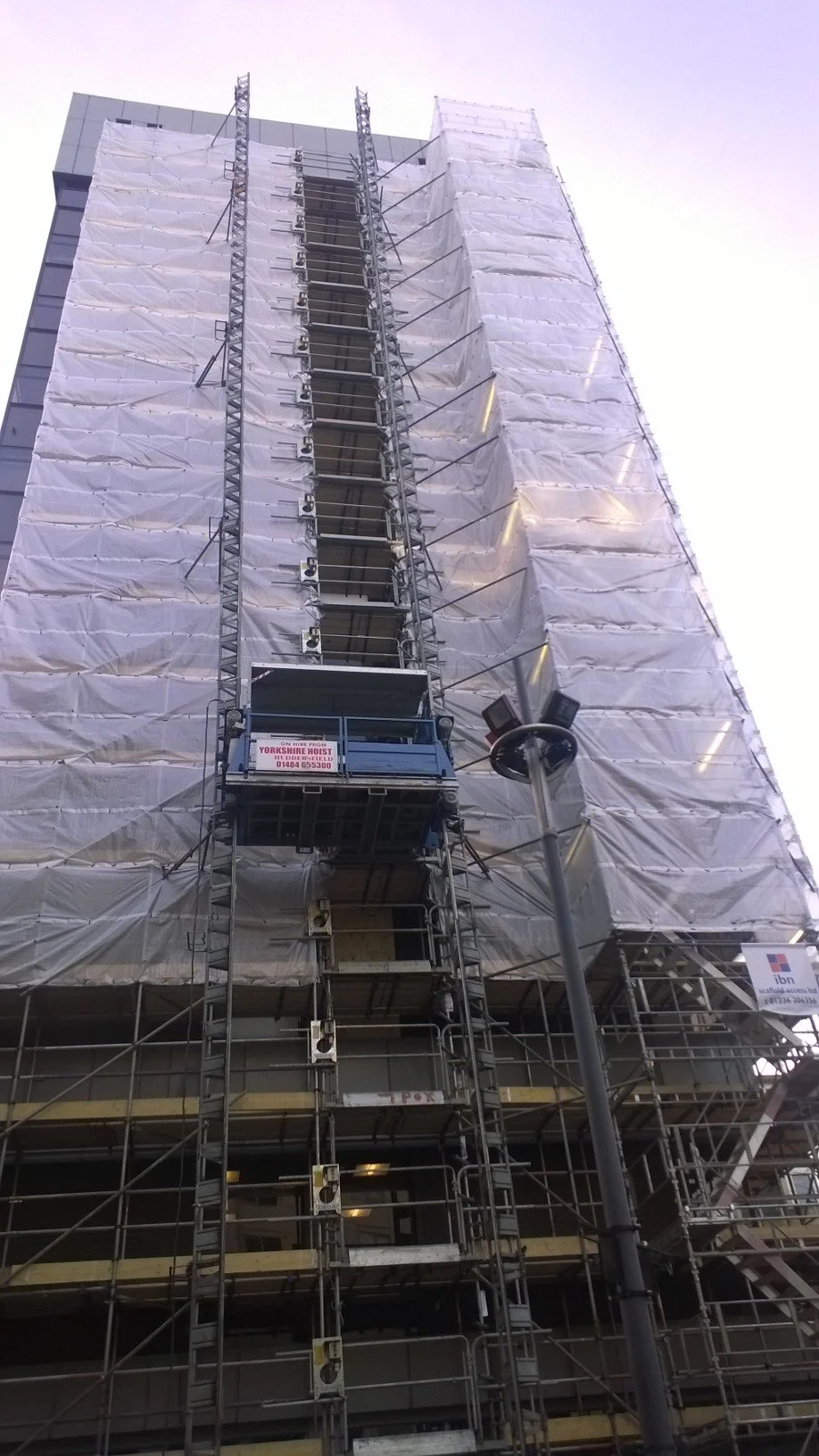 A tall building is covered in scaffolding and white protective sheeting for construction or renovation. A platform with a sign is attached to the scaffolding, and there is a street lamp with cameras nearby.