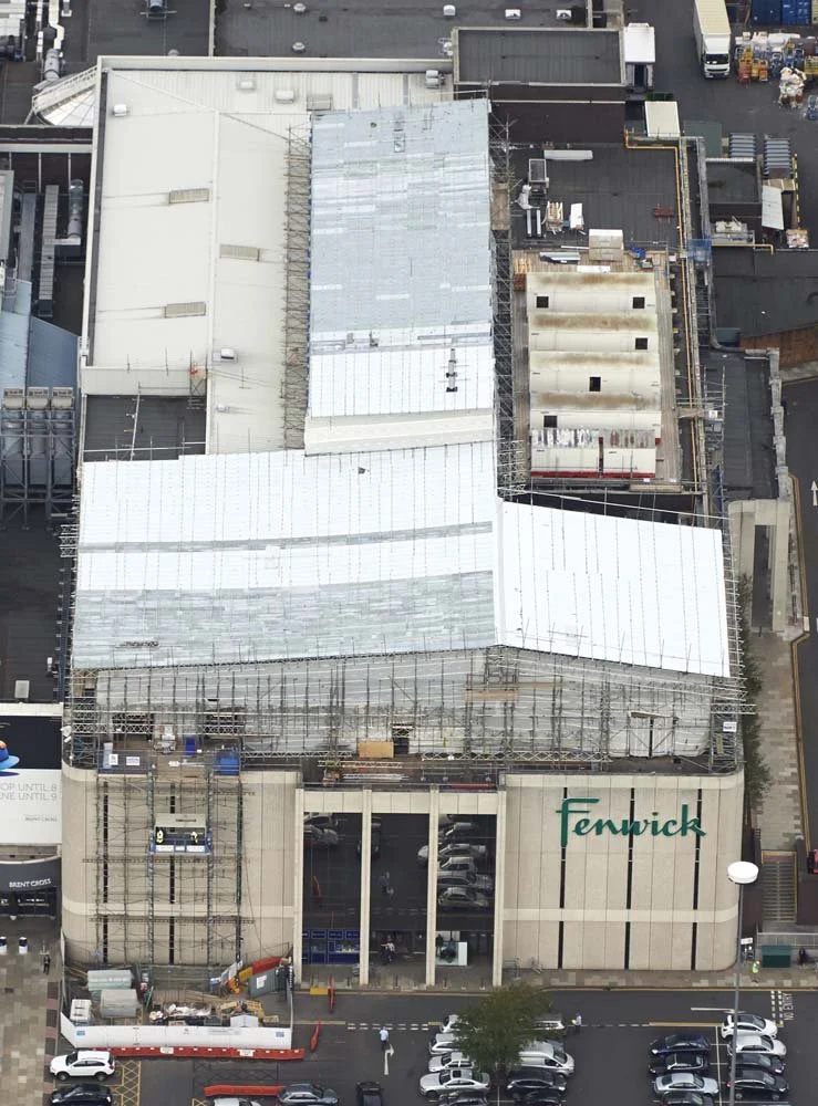 An aerial view of a shopping mall named Fenwick with construction scaffolding on its roof, indicating renovation work, and a parking lot with cars and pedestrians in front.