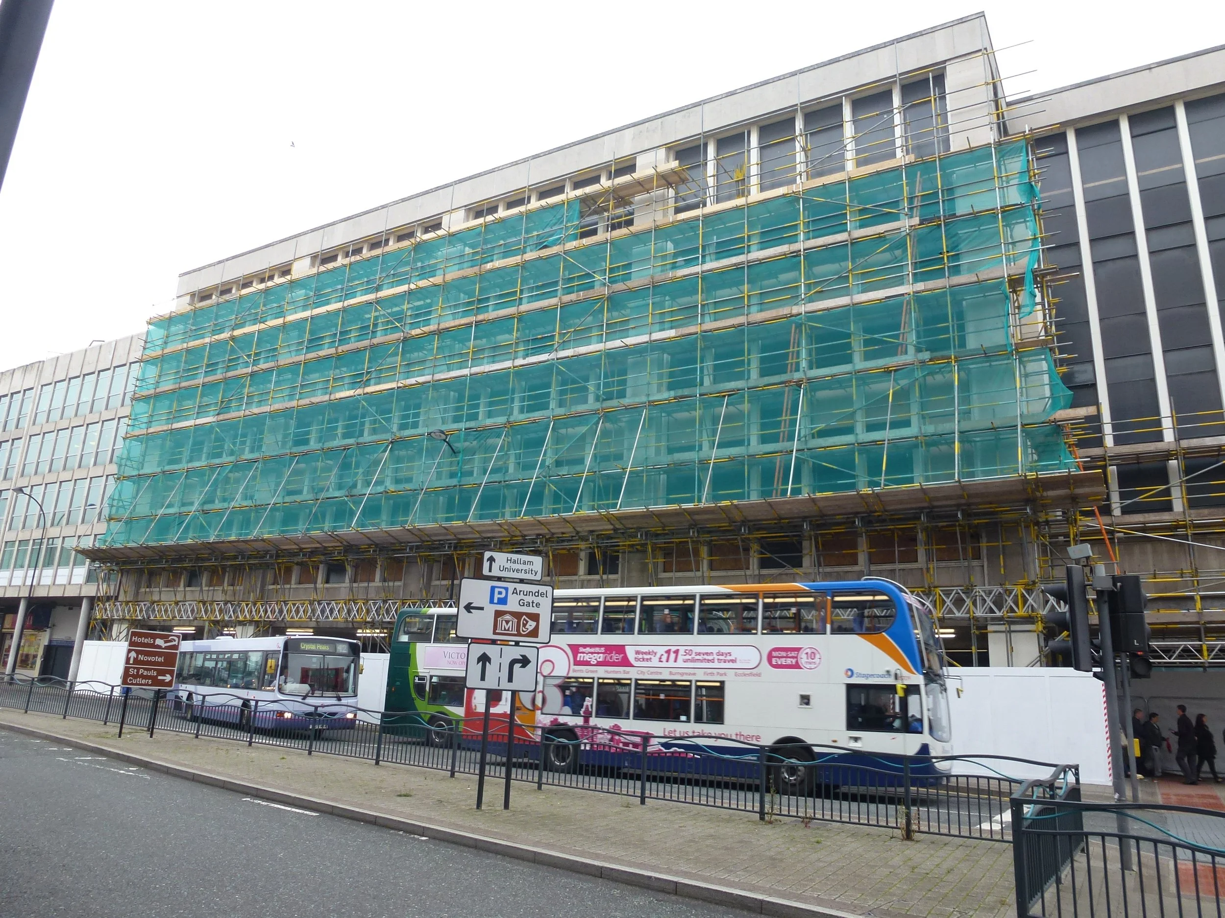 A building under construction with blue safety netting, several buses parked in front, and street signs indicating directions to hotels and parking.