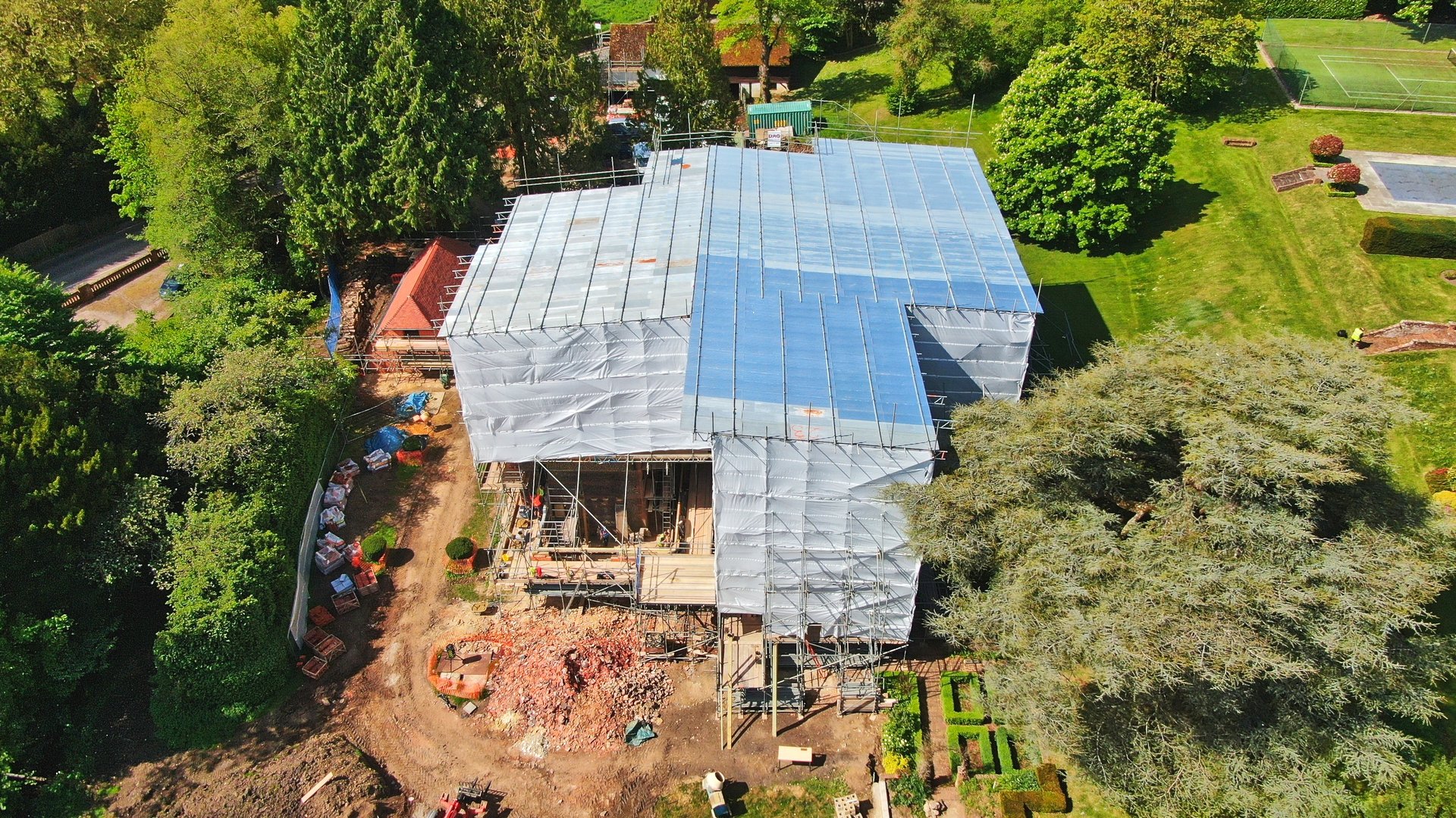 An aerial view of a house under construction with scaffolding and a partially installed roof, surrounded by trees and a well-maintained lawn with tennis courts in the background.