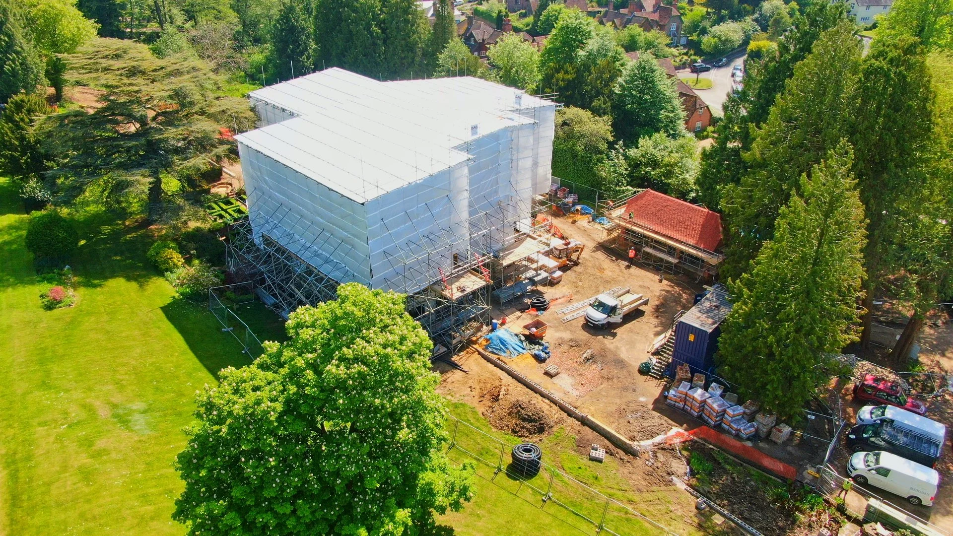 Aerial view of a construction site in a suburban area with green trees and landscaped yards. A large building is under renovation or construction, covered in scaffolding and white sheeting. Surrounding the site are construction materials, trucks, and