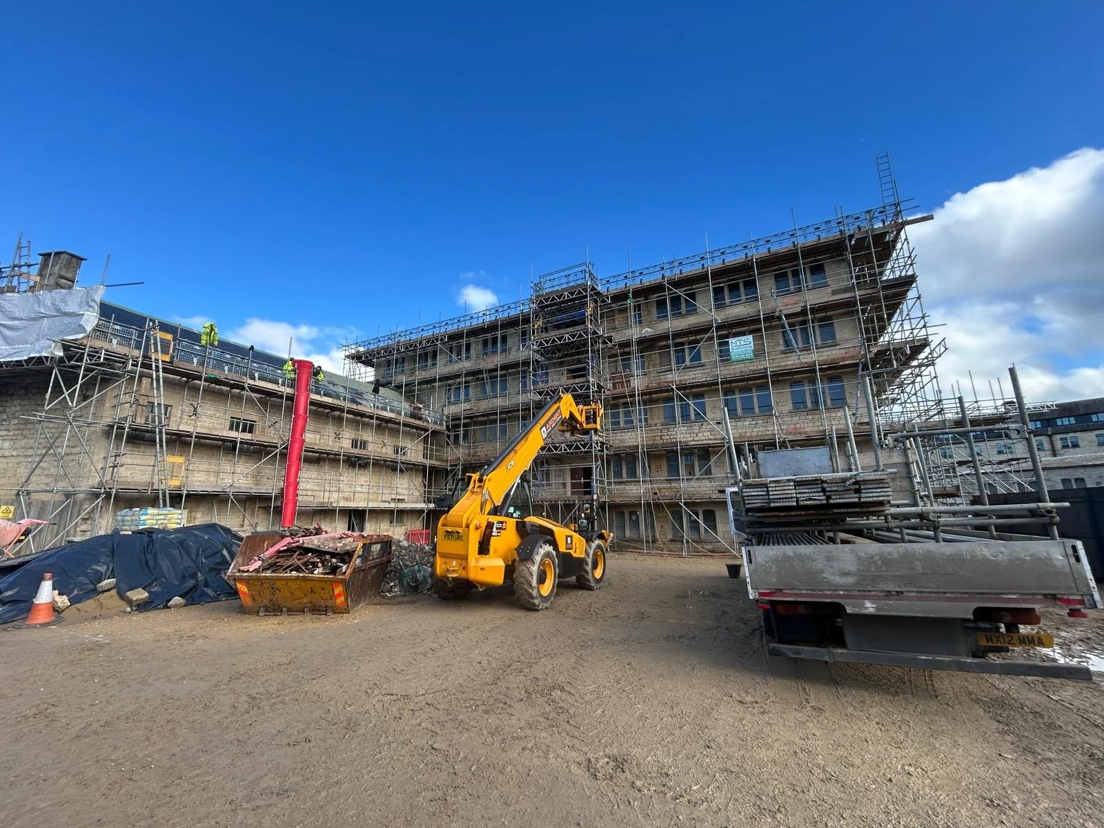 Construction site with scaffolding around a multi-story building, a yellow telescopic forklift, and a truck loaded with construction materials under a blue sky with some clouds.