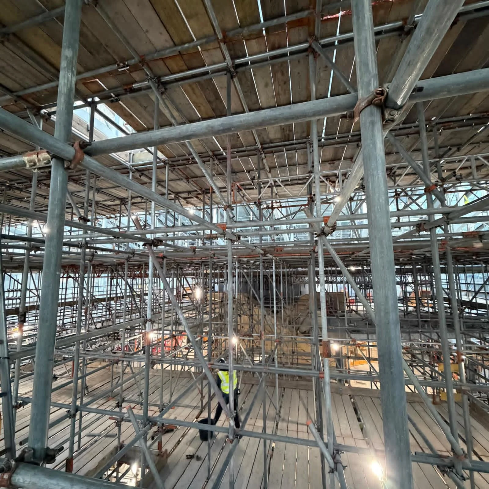 Construction site with a complex network of metal scaffolding and a worker wearing a yellow safety vest.
