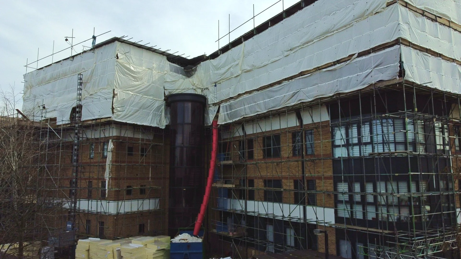 Building under construction with scaffolding and white protective cover, brick facade, and black cylindrical structure with red pipe.