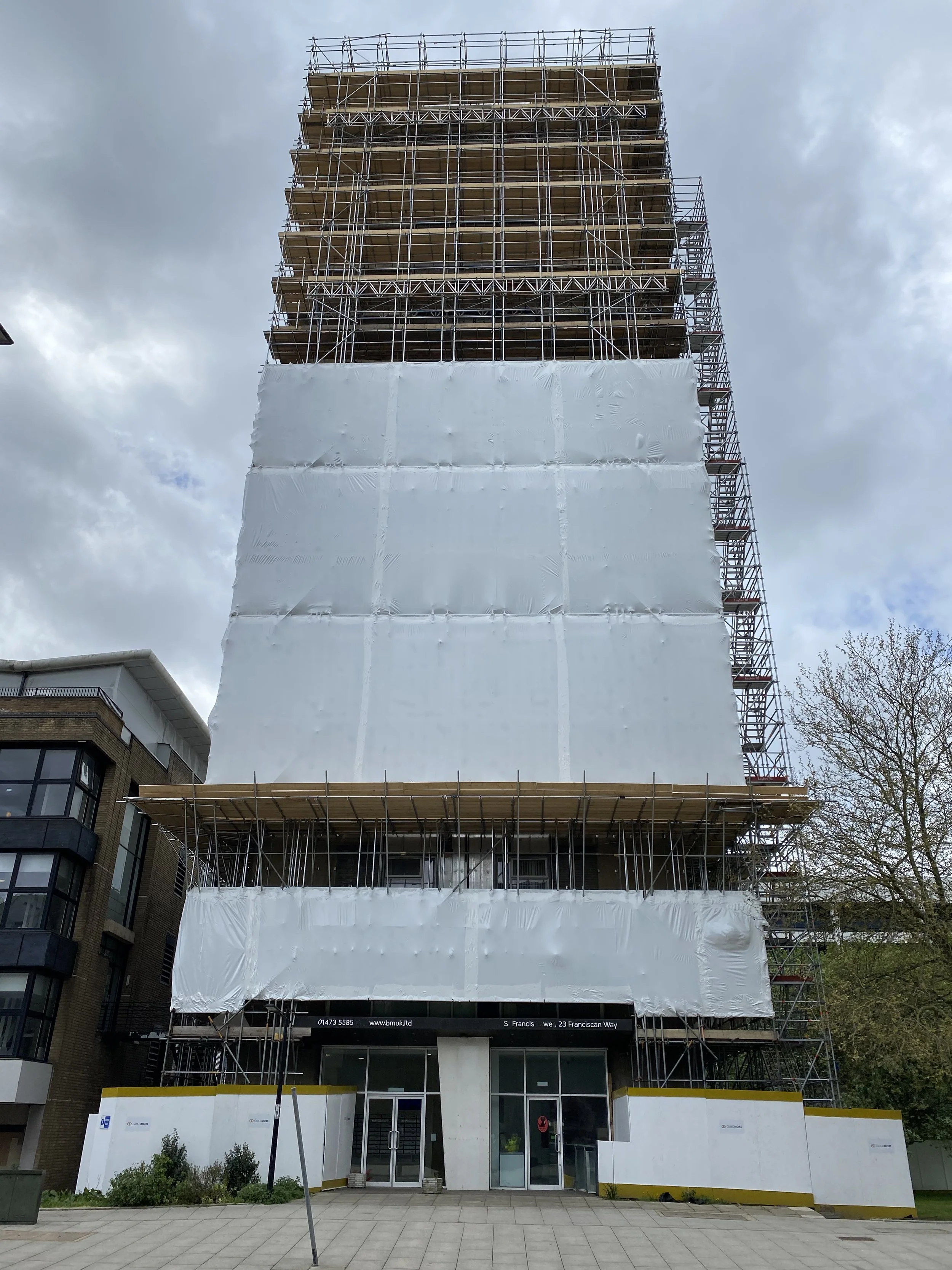 A tall building under construction covered in scaffolding, with white protective wrap on part of the structure, and an entrance below with glass doors.