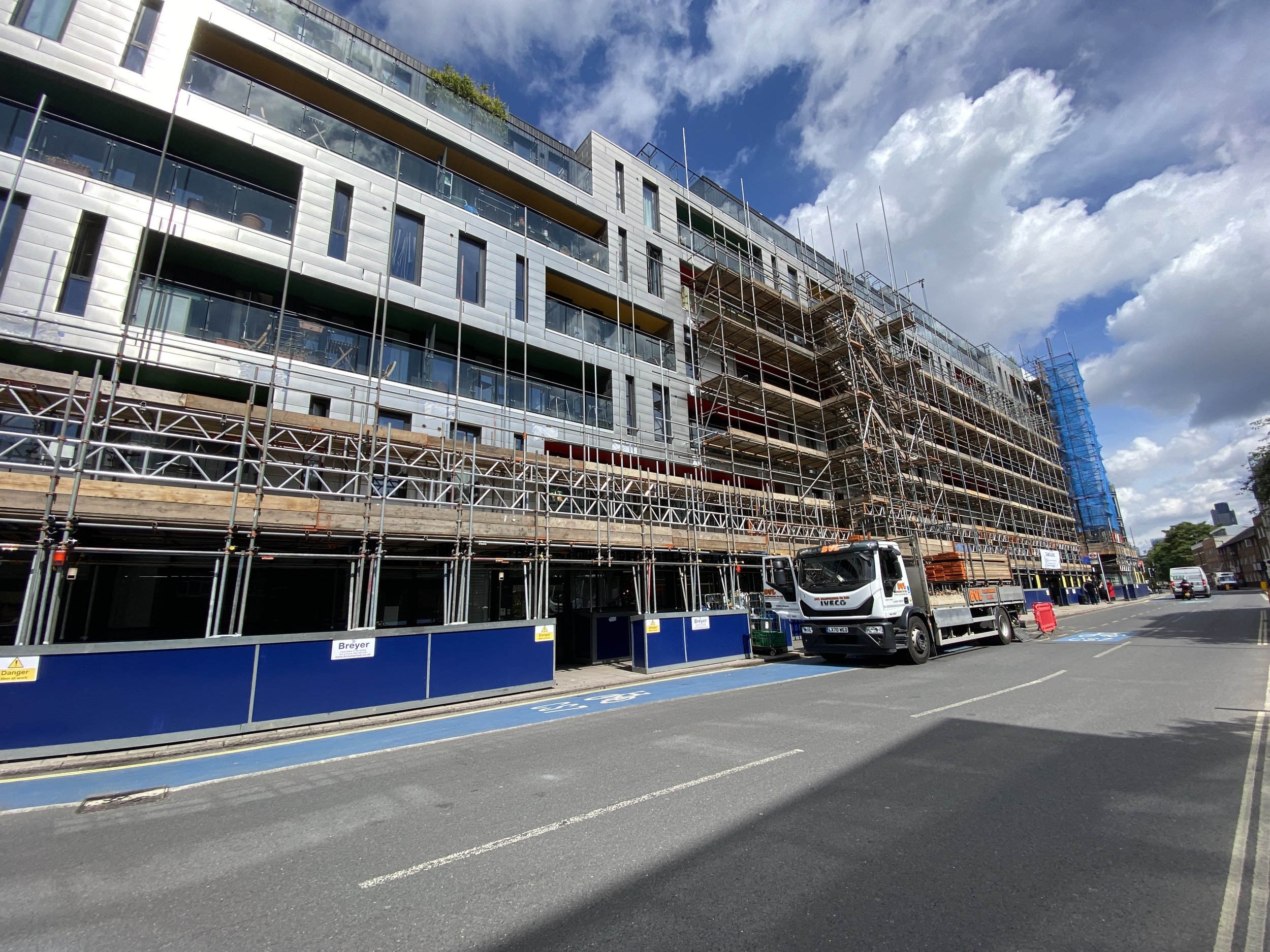 Construction site with scaffolding surrounding a modern multi-story building under development, parked trucks, and a partly cloudy sky.