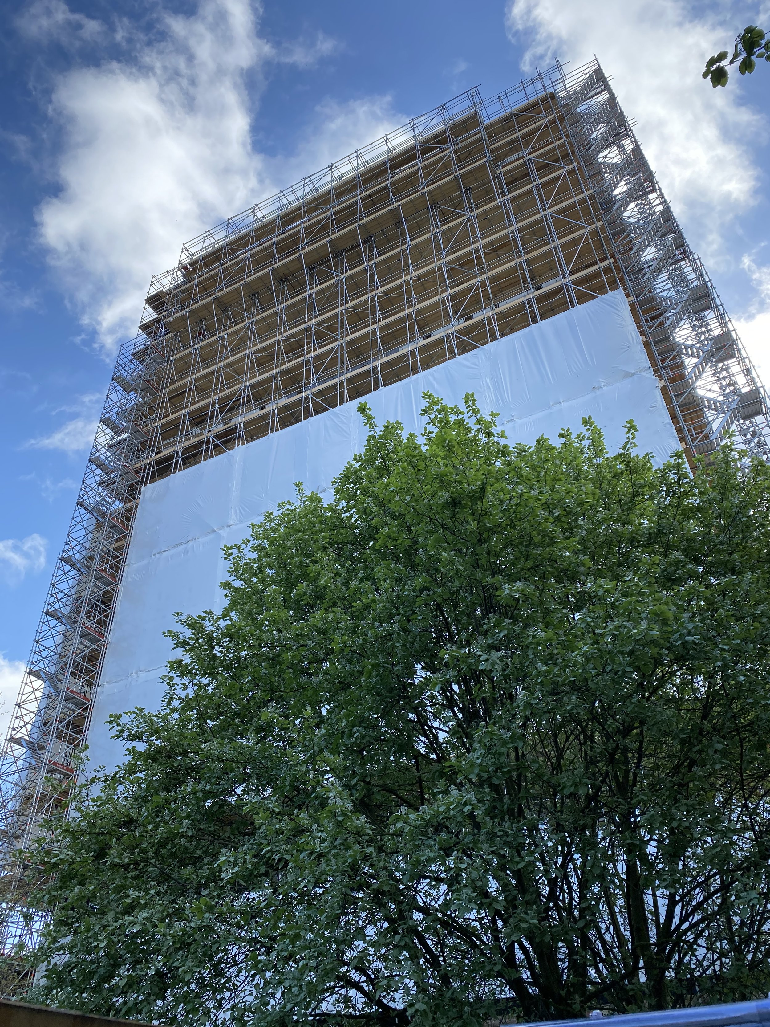 A high-rise building under construction with scaffolding around it and white protective covering on the lower part, with a green tree in the foreground and a partly cloudy blue sky in the background.
