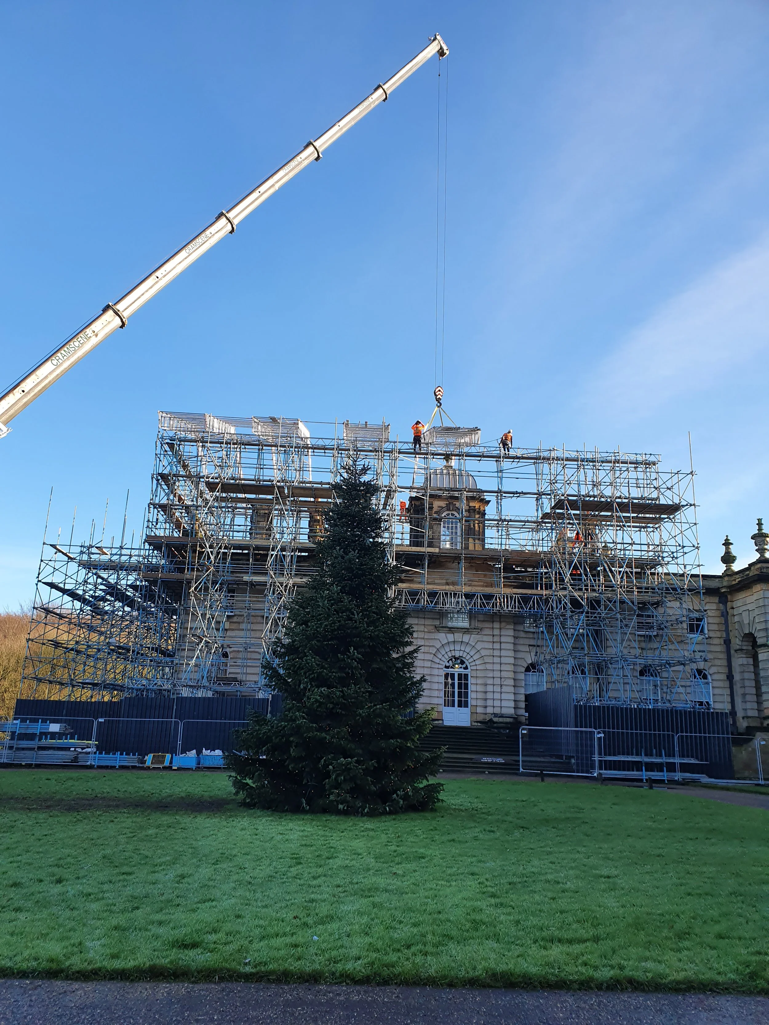 Construction workers and scaffolding surrounding a historic building, with a crane lifting materials, a Christmas tree in the foreground, and a blue sky.