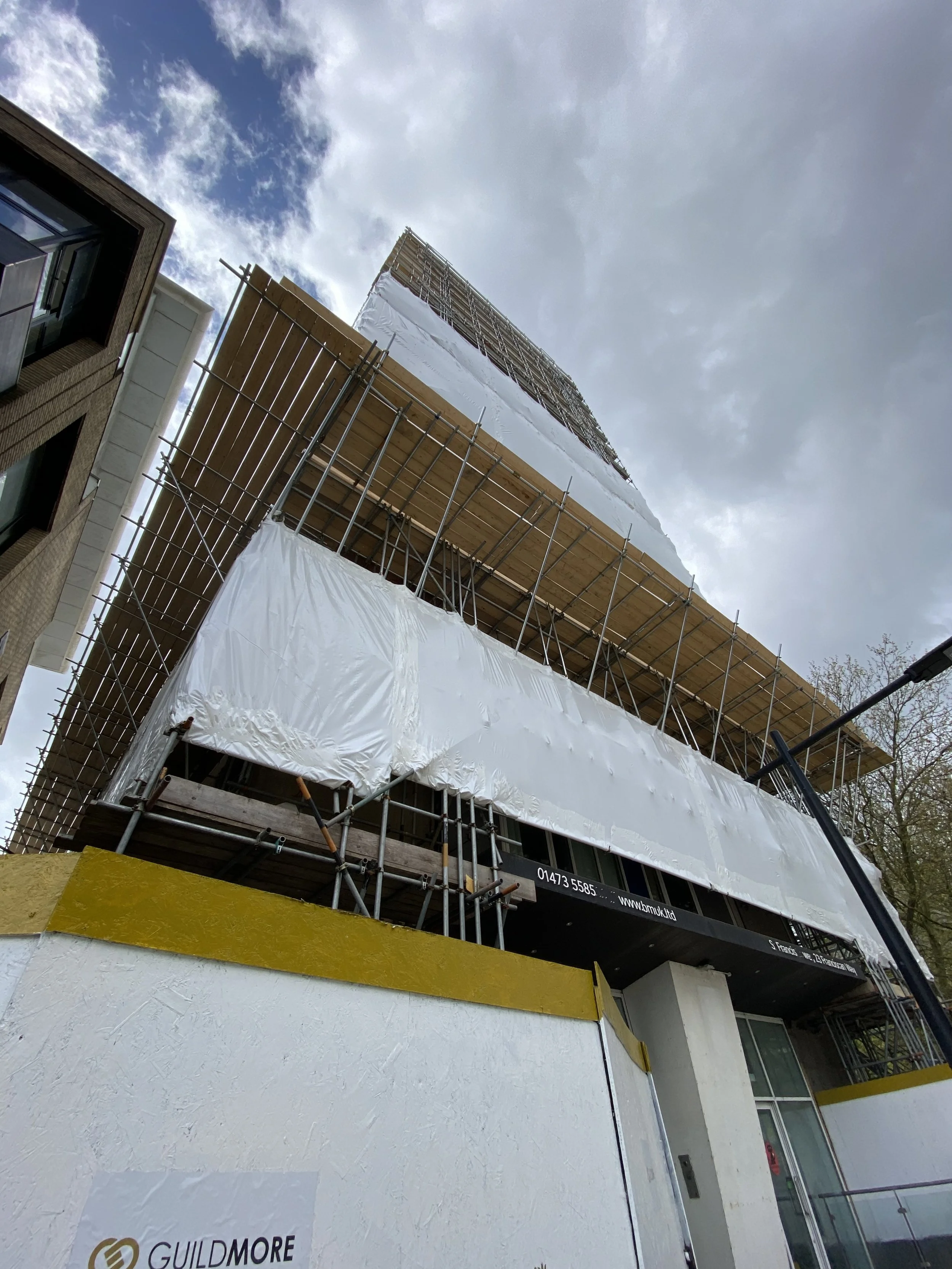 A multi-story building under construction with scaffolding and protective white plastic sheeting, viewed from a low angle against a cloudy sky.