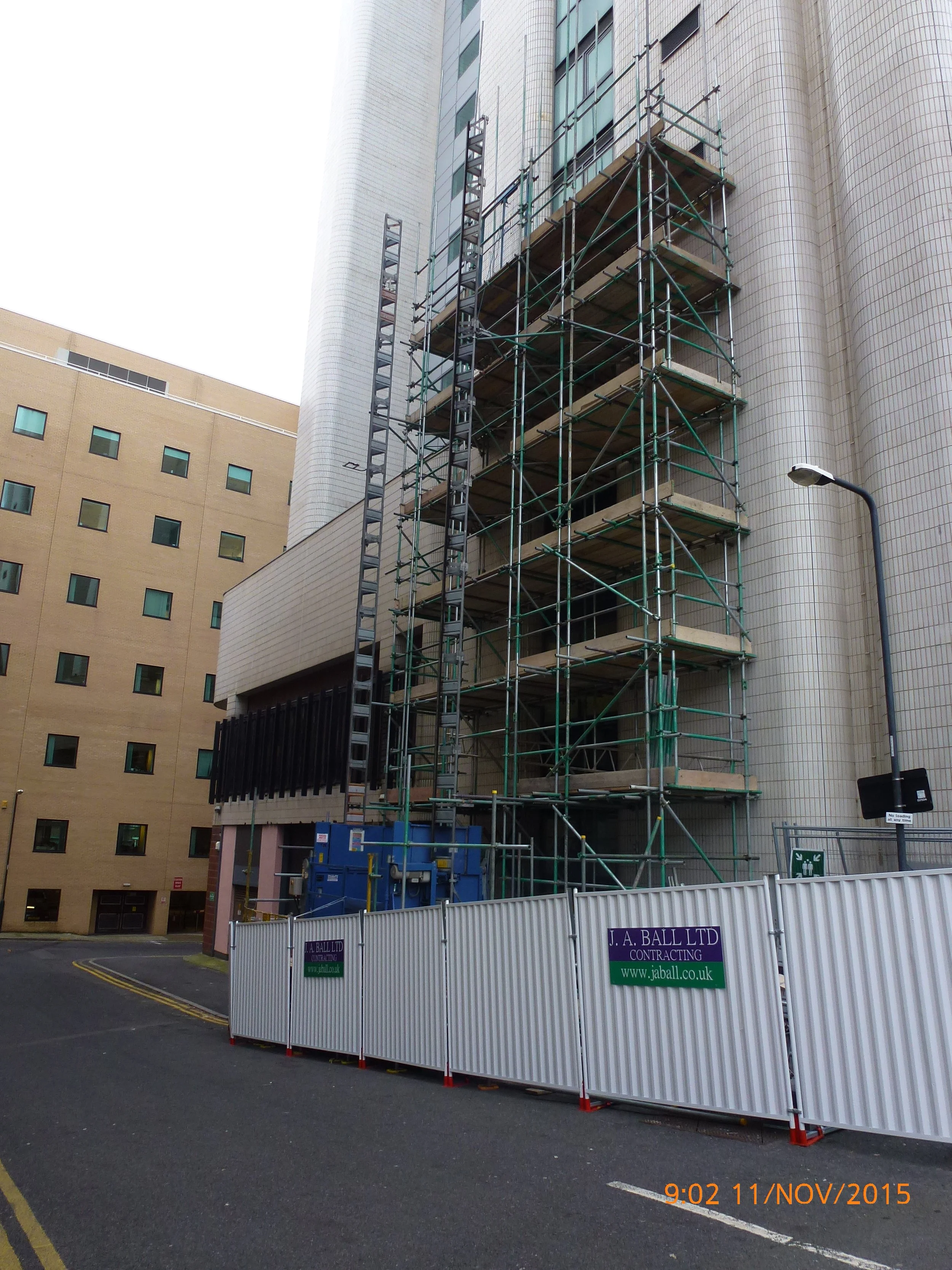 Construction site with scaffolding surrounding a multi-story building in an urban area, with a white safety barrier and a sign for J. A. Ball Ltd Contracting.