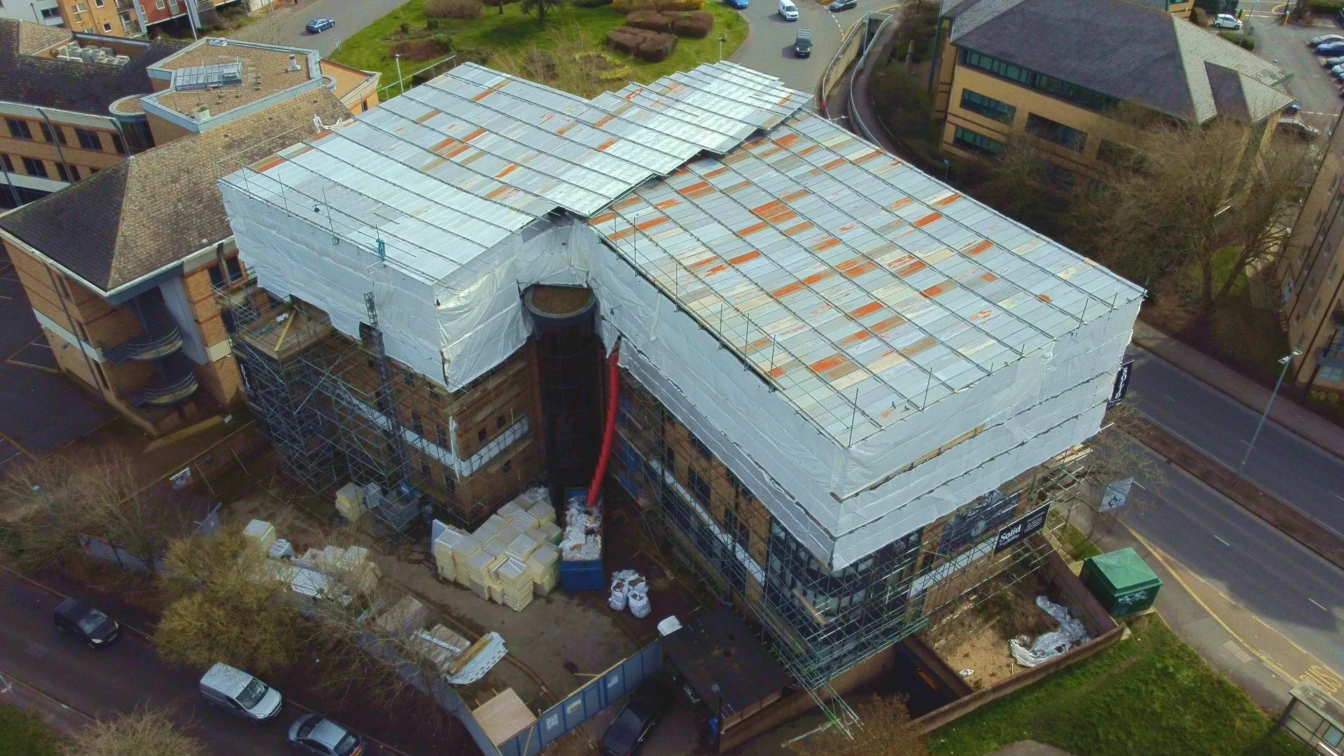 An aerial view of a building under renovation with scaffolding and a partially covered roof, surrounded by parked cars and nearby buildings.