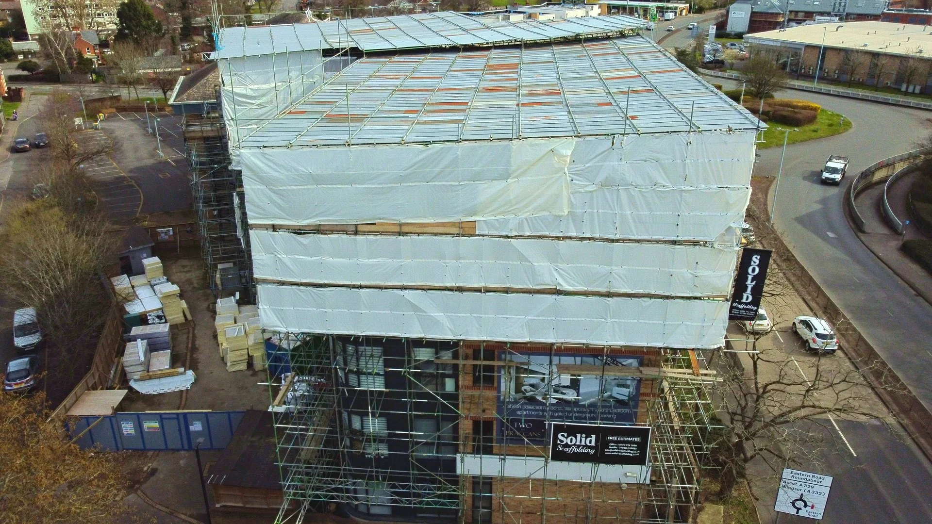 A building under construction with scaffolding and white protective sheeting, next to a parking lot and a curved road.