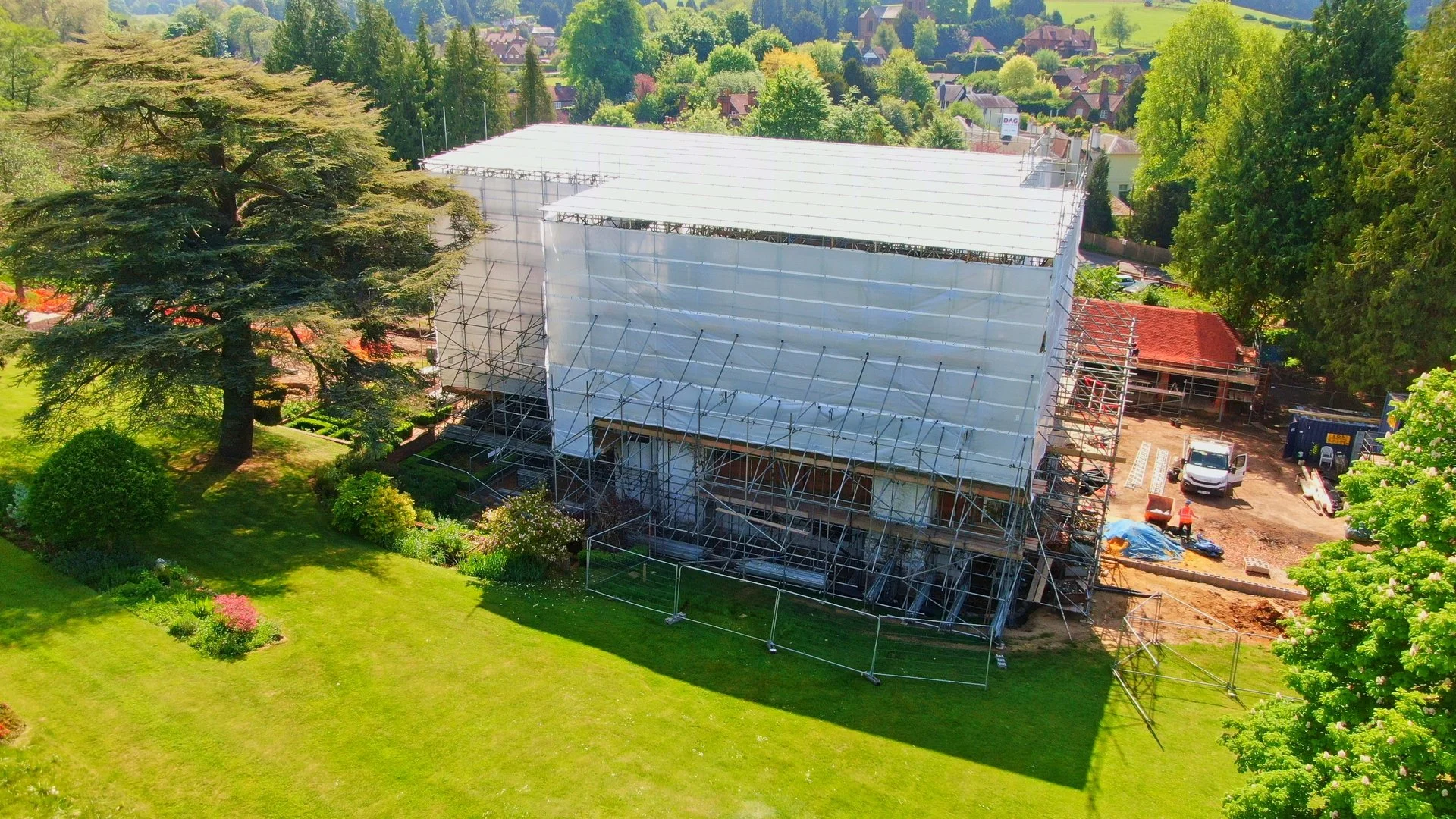 A house under construction surrounded by a lush green garden with large trees and flowering plants. Construction scaffolding and equipment are visible around the building.