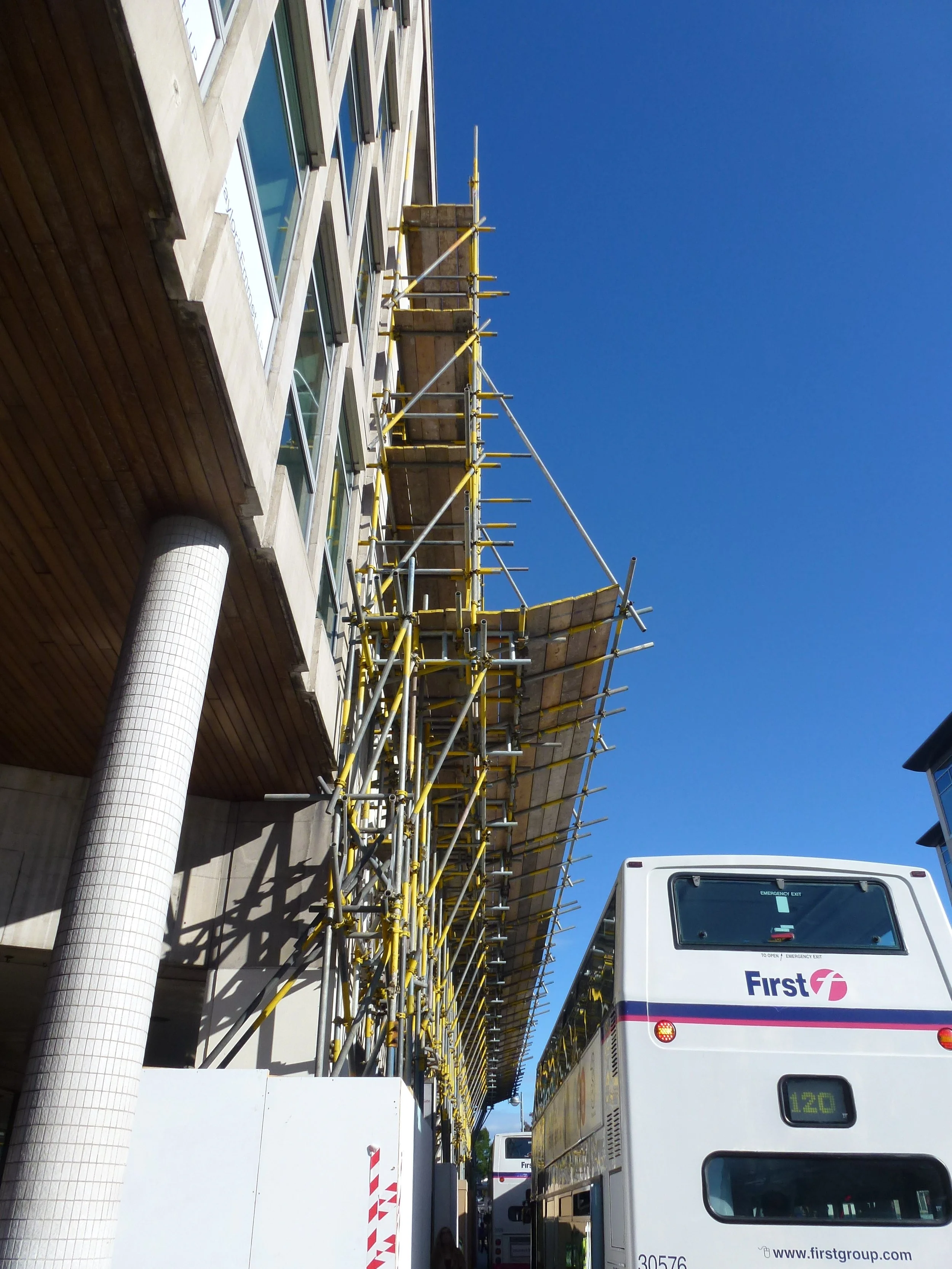 Construction scaffolding installed along the side of a multi-story building with large windows, with a white city bus parked nearby on a bright, clear day.
