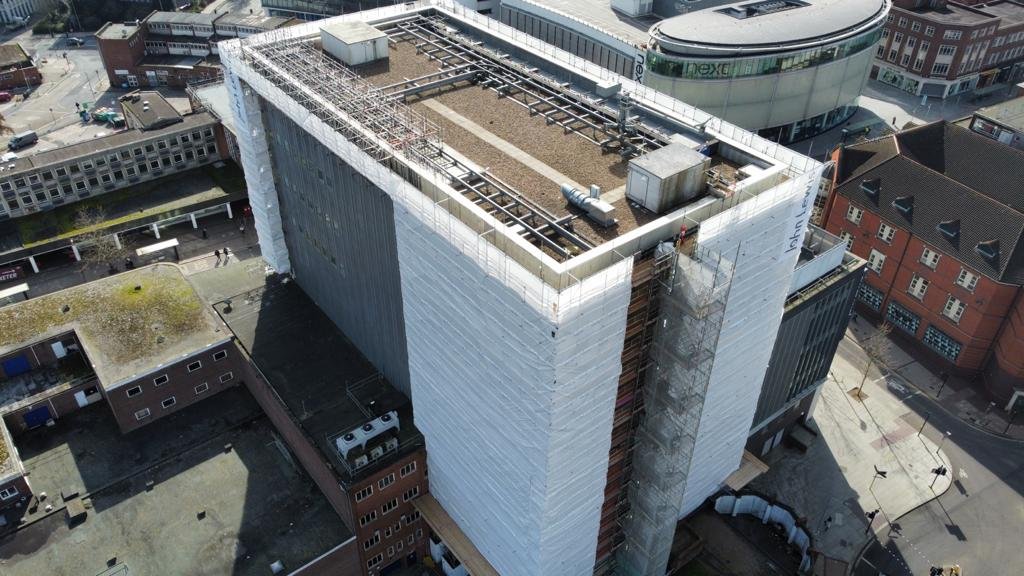 Aerial view of a multi-story building under construction with scaffolding and construction equipment on the rooftop, surrounded by other buildings.