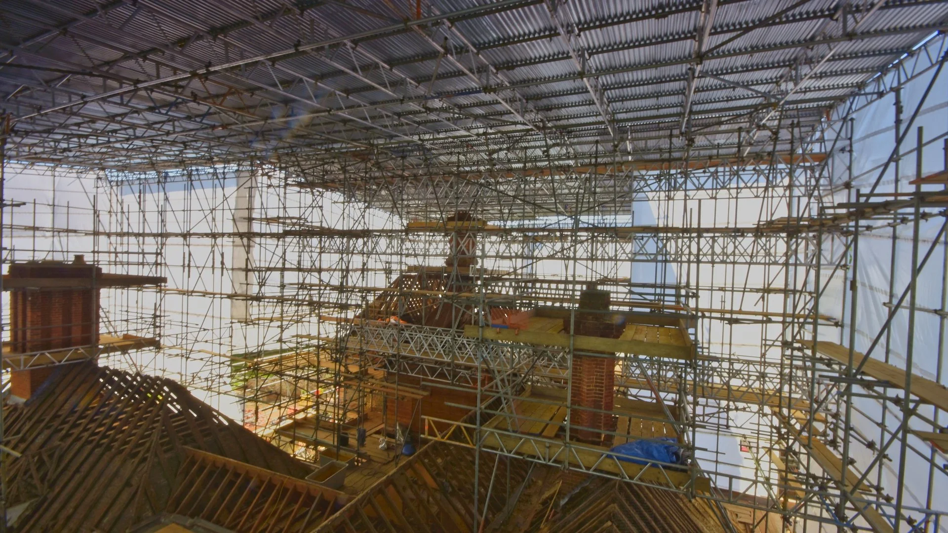 An indoor construction site with extensive metal scaffolding and partially visible brick chimney stacks, indicating ongoing renovation of a building's roof structure.