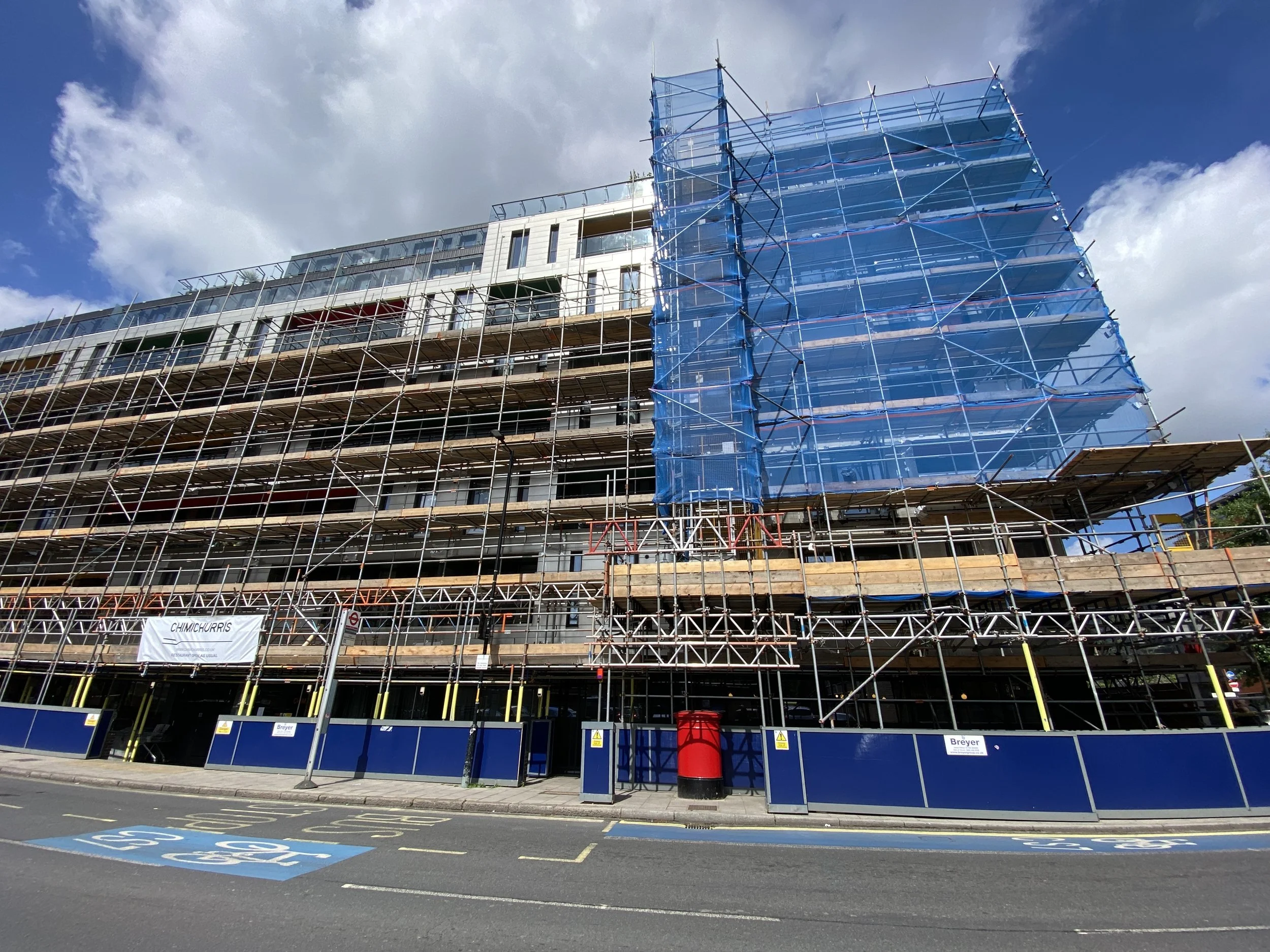 Construction site with scaffolding surrounding a multi-story building, some covered with blue netting, in an urban area under a partly cloudy sky.