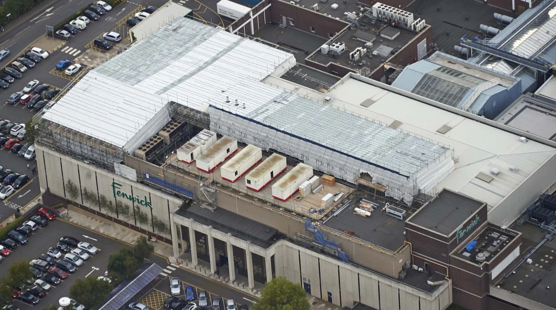 Aerial view of Fenwick department store with construction work on the roof, surrounded by parking lots and nearby buildings.