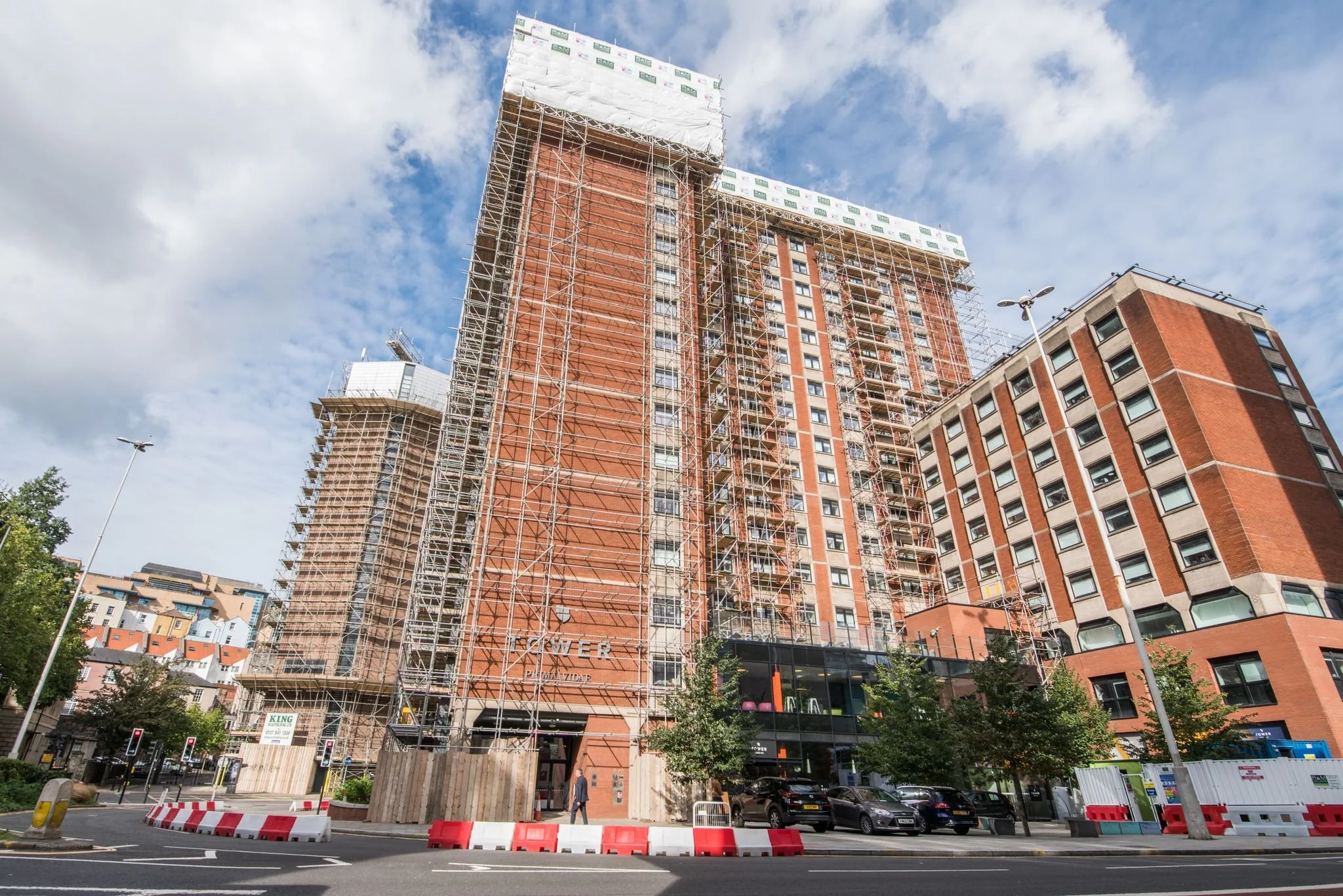 Under construction high-rise building with scaffolding on a city street with cars and trees.