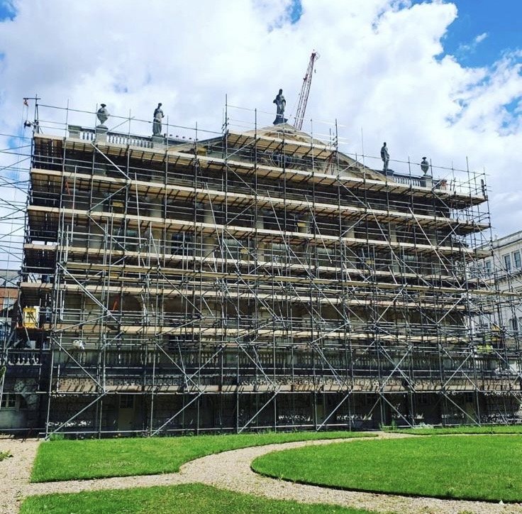 Historic building under renovation with scaffolding, featuring statues on top and a crane in the background.