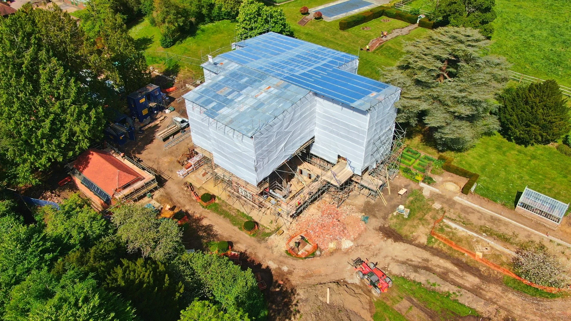 An aerial view of a construction site with a house under renovation, covered in white and transparent plastic sheeting, surrounded by green trees and lawns.
