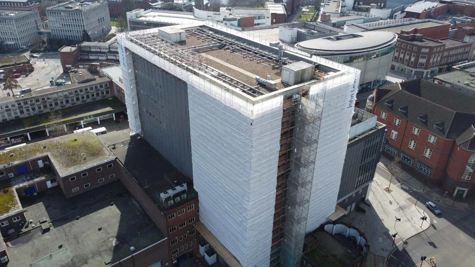 Aerial view of a high-rise building under construction in an urban area, with scaffolding wrapped around parts of the building and surrounding streets with parked cars and smaller buildings.