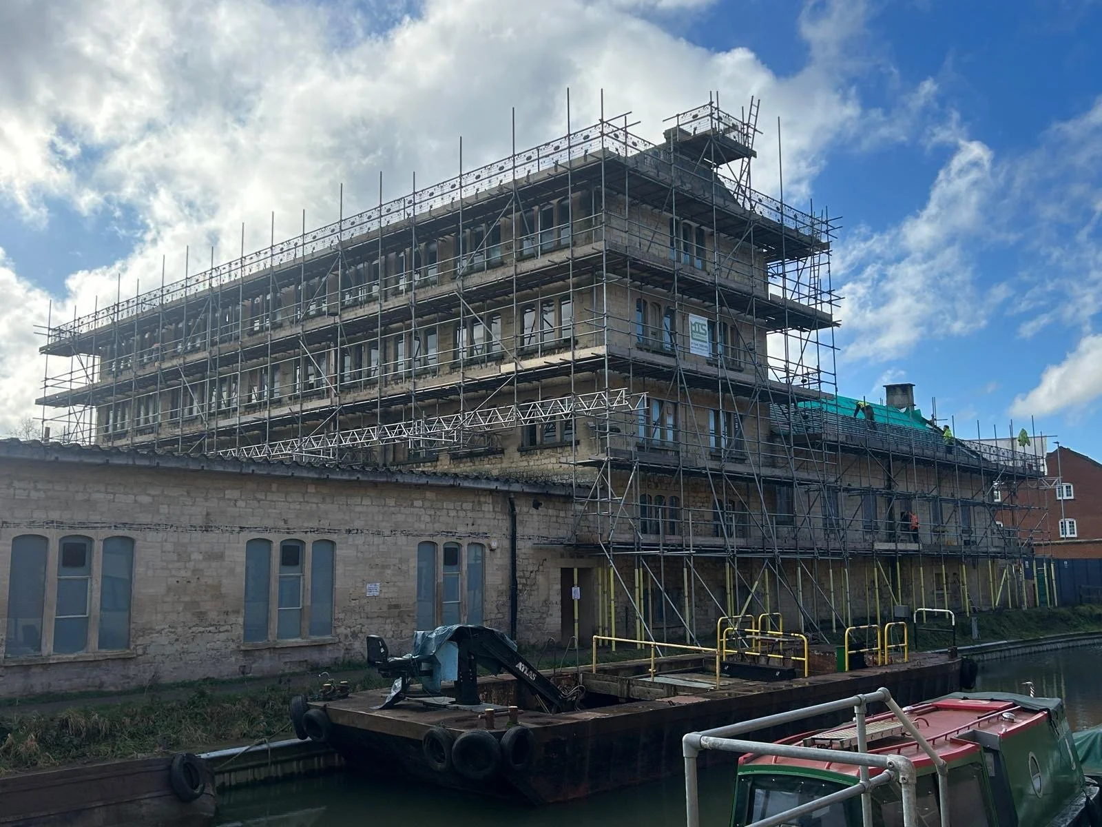 A multi-story building under construction with scaffolding around it, located beside a waterway, with a boat docked at the water's edge, and a partly cloudy sky overhead.
