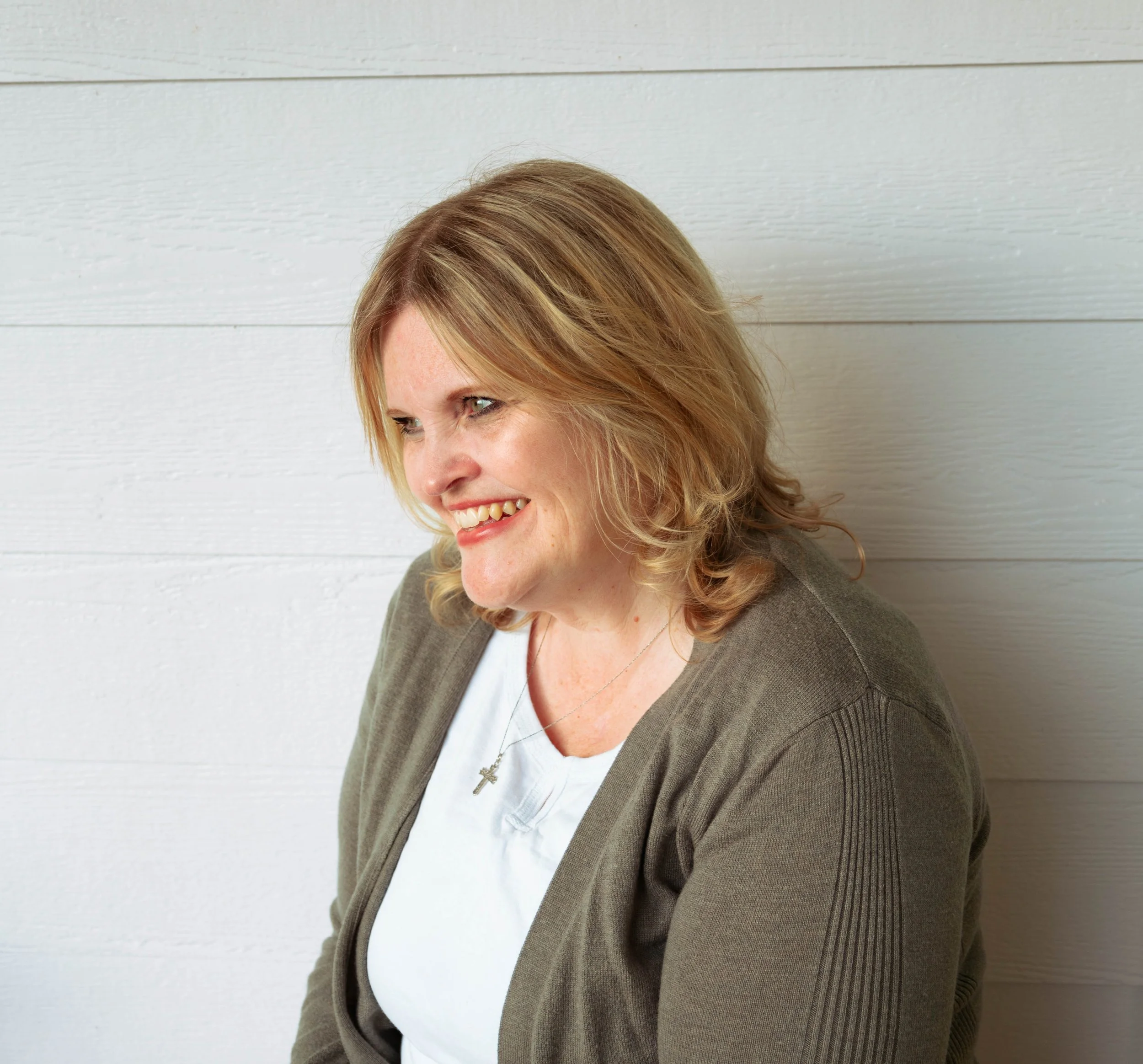 Close-up of a smiling woman with shoulder-length blonde hair, wearing a gray blazer over a white top with a cross necklace, standing against a light-colored wooden wall.