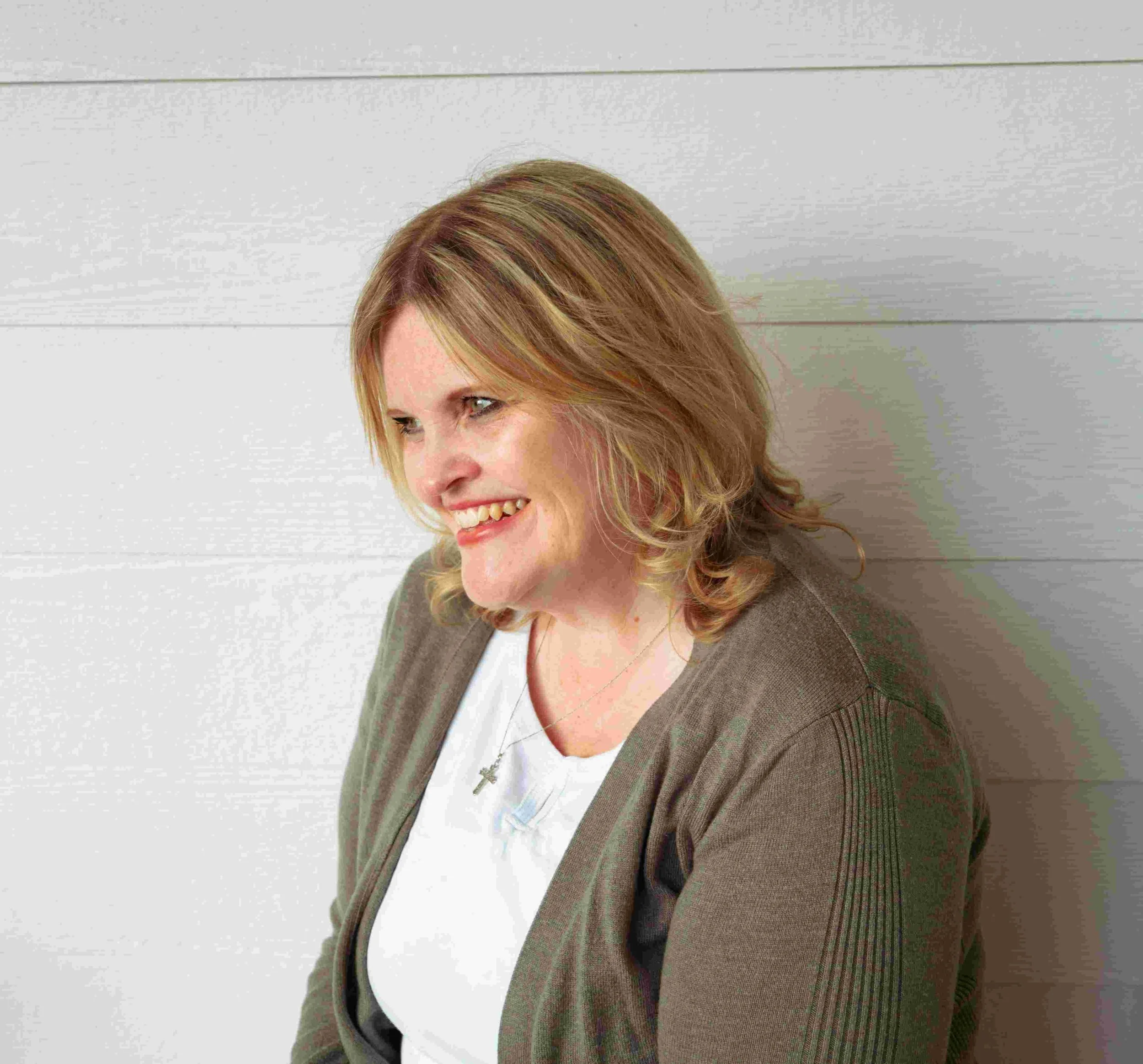 Smiling woman with blonde hair wearing a brown cardigan and cross necklace in front of a white wooden wall
