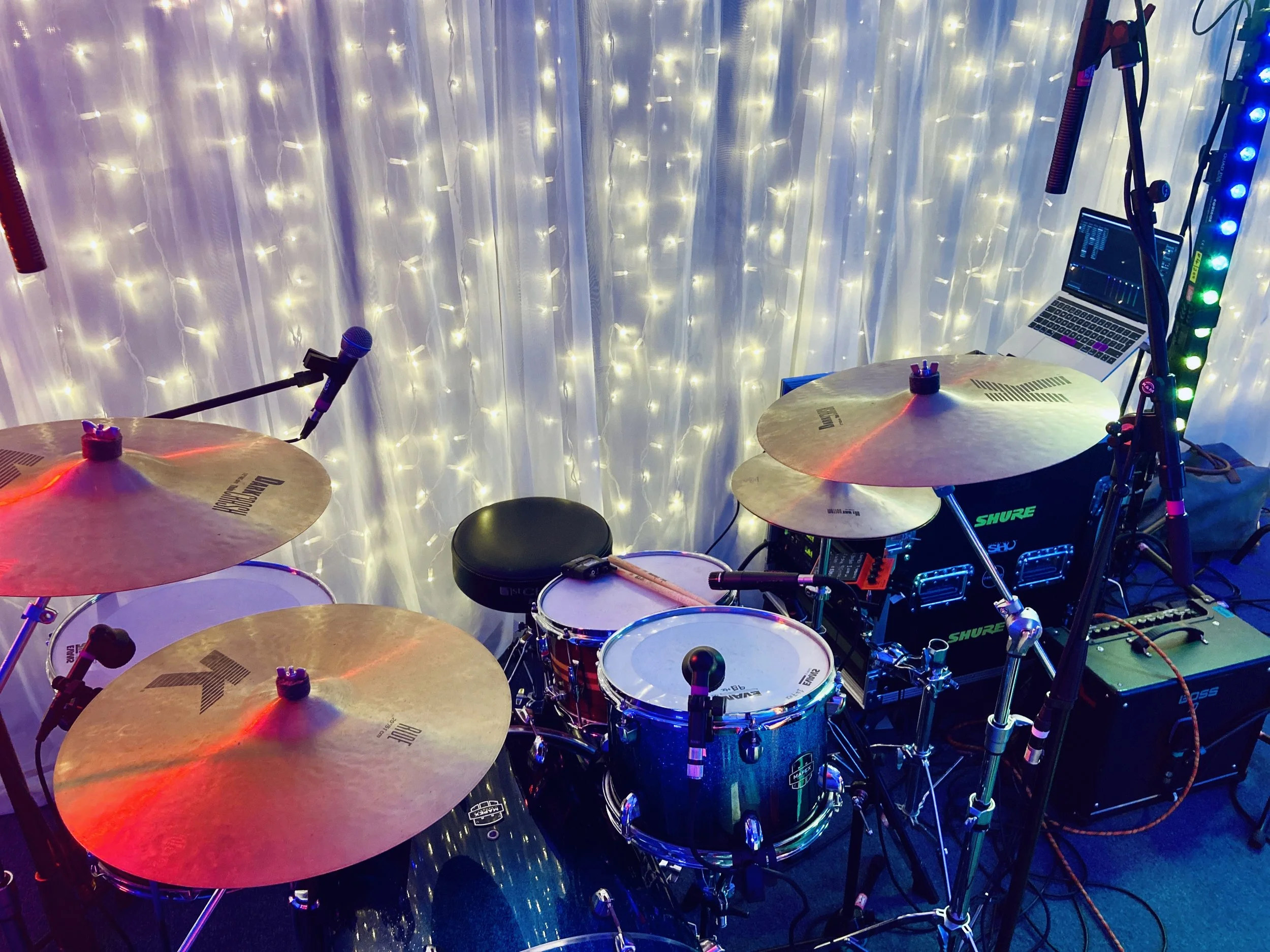 Music setup with a drum kit, microphones, and electronic equipment against a backdrop of white curtains with string lights.