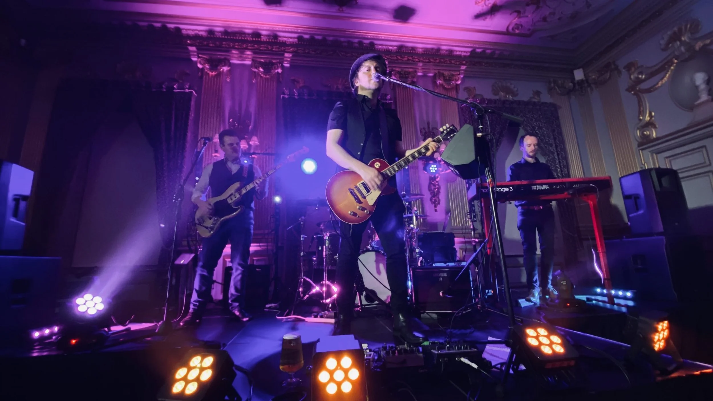 A band performing live on a stage with purple and orange lighting, featuring a female guitarist and vocalist, a male guitarist, and a male keyboardist in an ornate room.