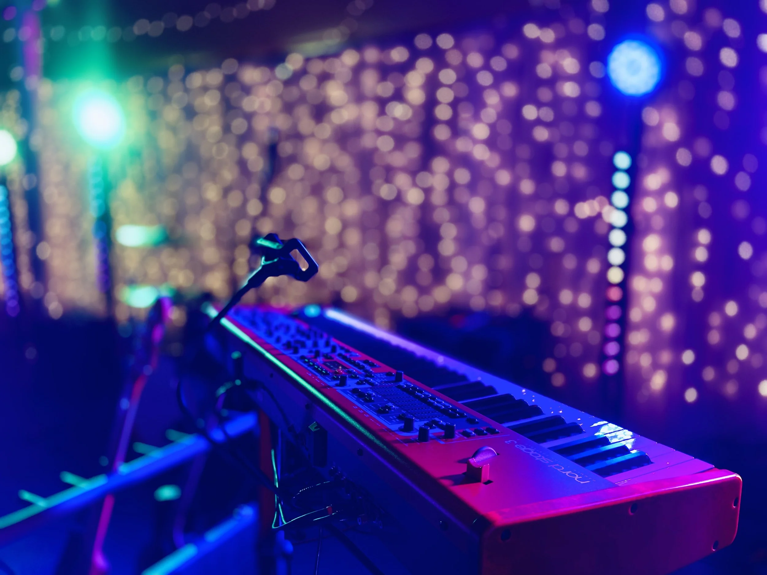 A colorful keyboard synthesizer on a stand, illuminated by vibrant pink, purple, and blue stage lights, with a blurred background of colorful bokeh lights.
