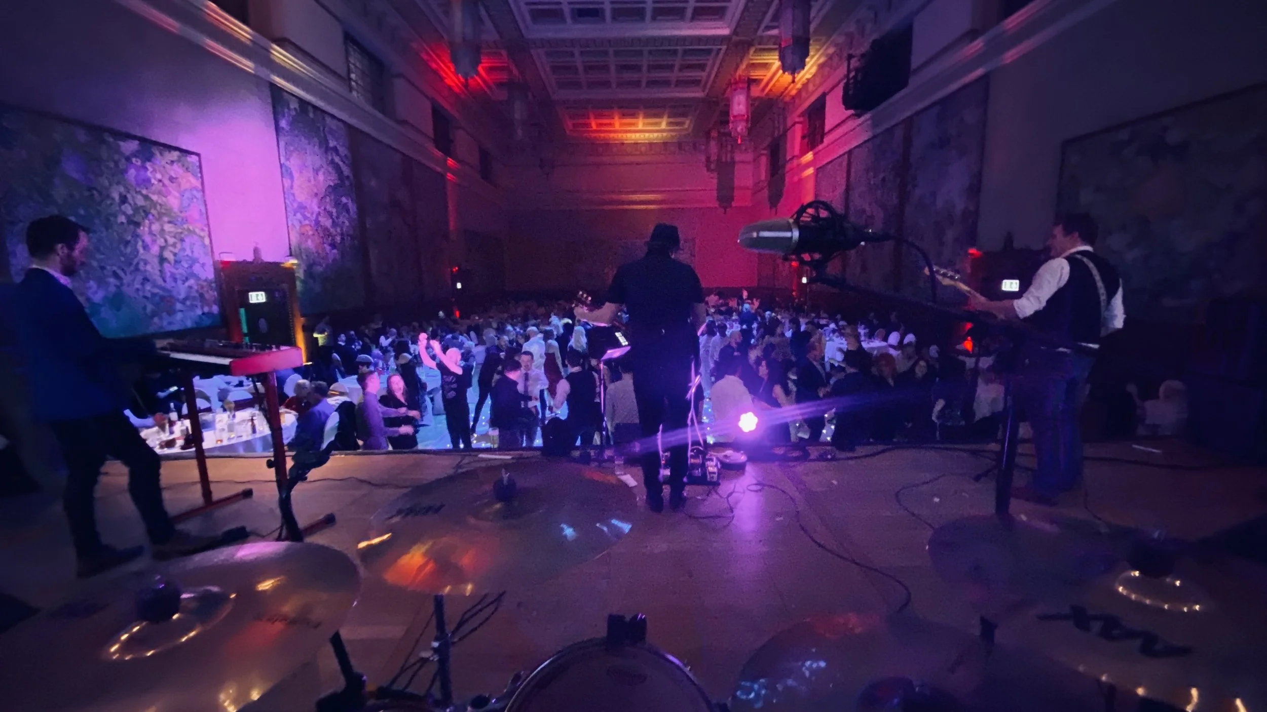 View of a band performing on stage at a large indoor event or dance party, with a crowded dance floor in front. The scene is illuminated with purple and pink stage lights, and the hall has large paintings or murals on the walls.