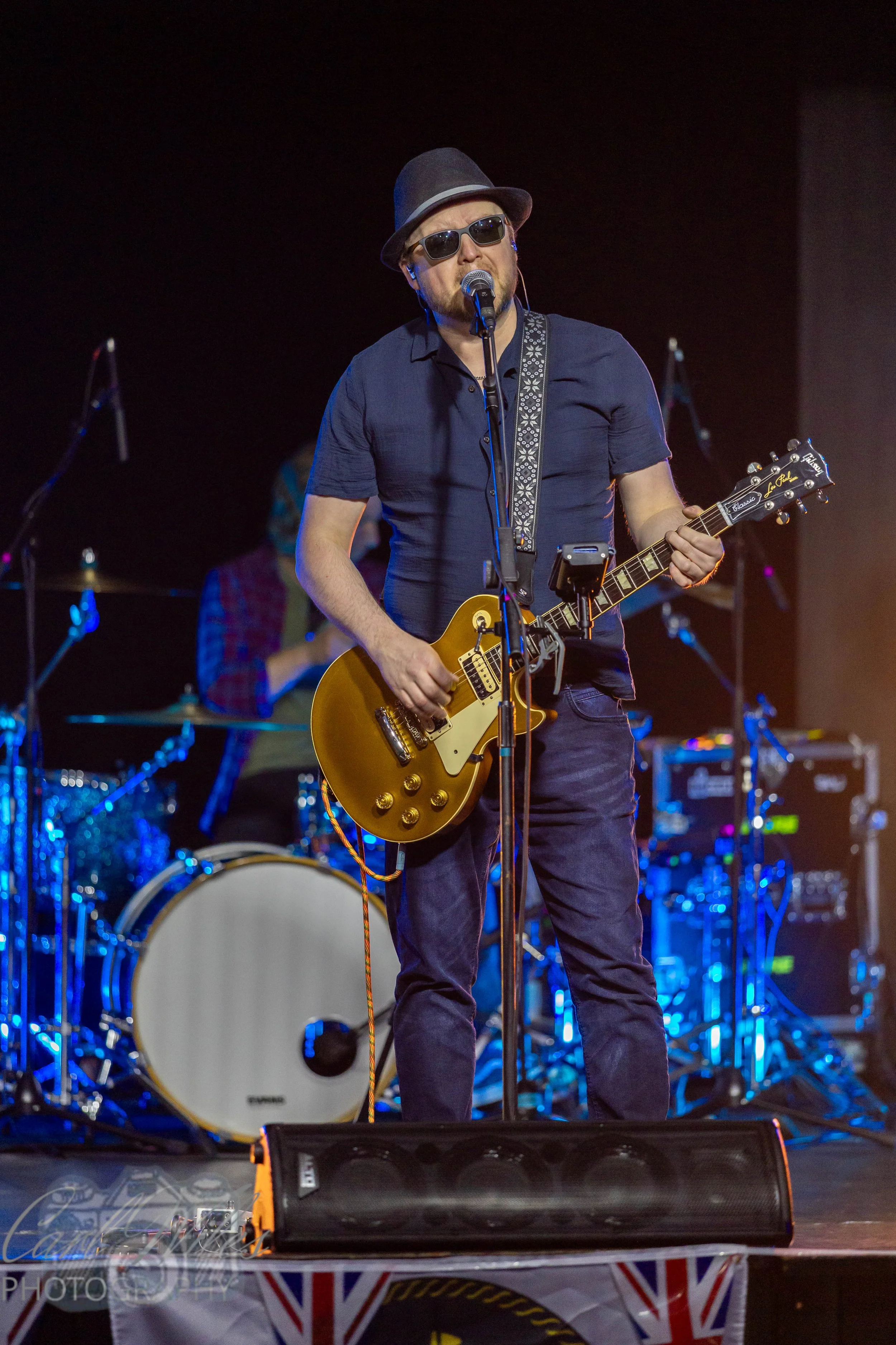 A man singing into a microphone on stage, wearing sunglasses, a fedora, and a dark shirt, playing an electric guitar. Behind him, there is a drummer and other band members, with stage lights illuminating the scene.